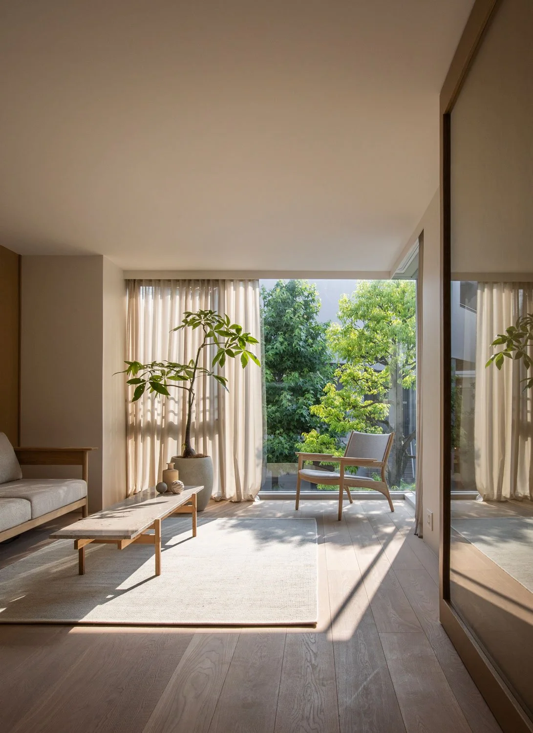 Living room with a large window showing green trees outside, a beige sofa, a small coffee table, a potted plant, two chairs, and curtains.