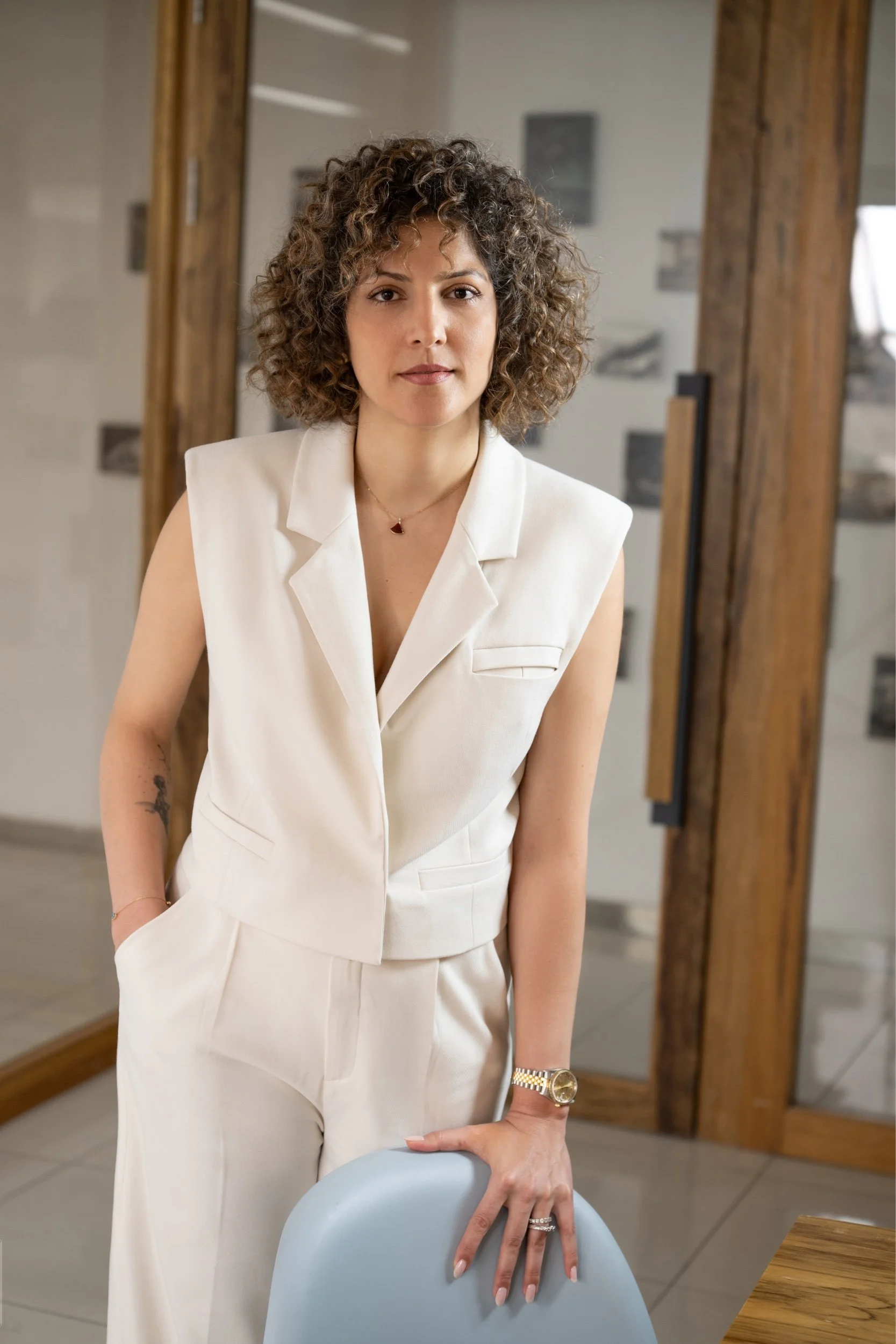 A woman with curly hair wearing a white sleeveless suit and gold watch, standing in a modern interior with wooden accents, resting her hand on a light blue chair.