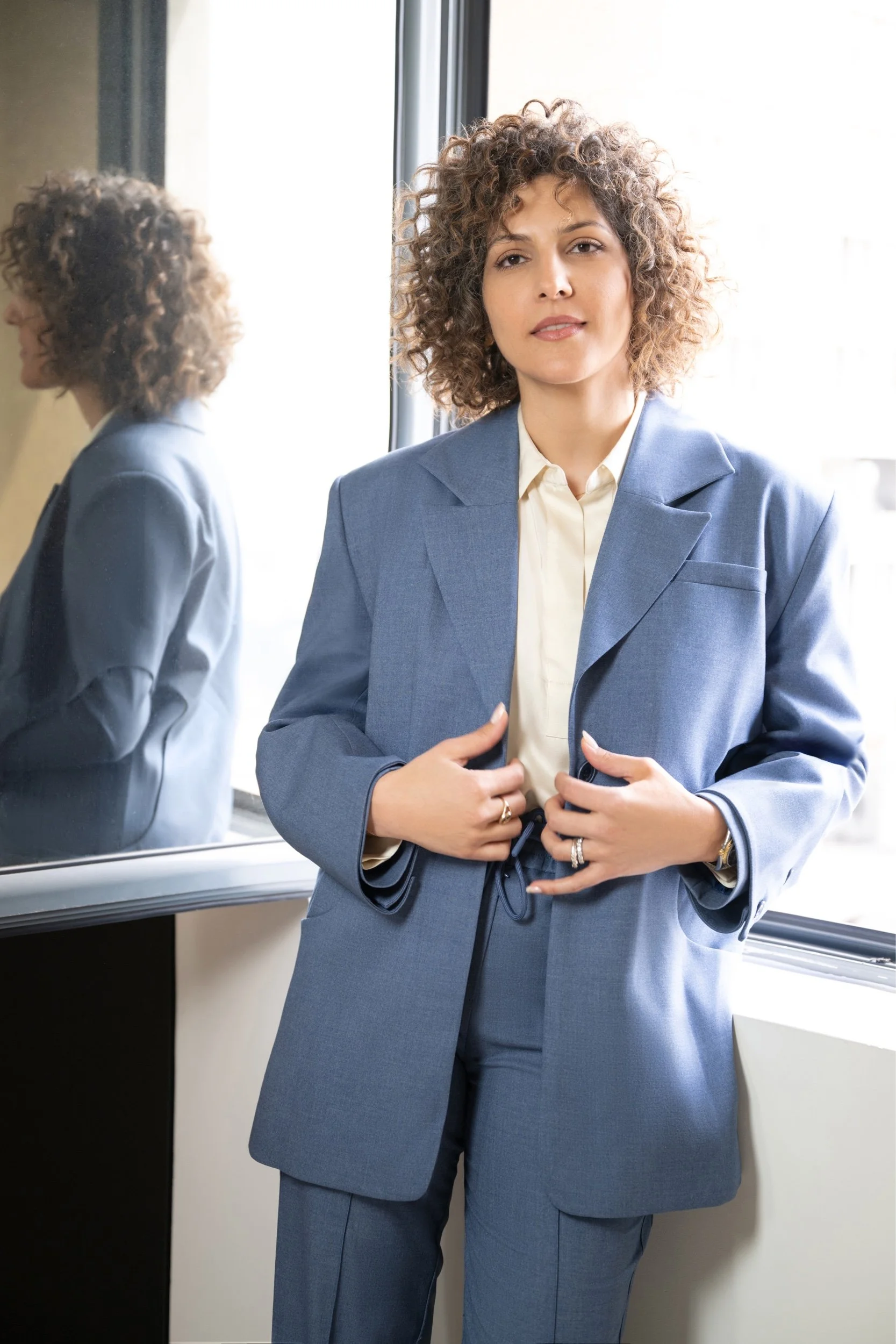 A woman with curly hair in a gray suit standing near a window, with her reflection visible in the mirror behind her.