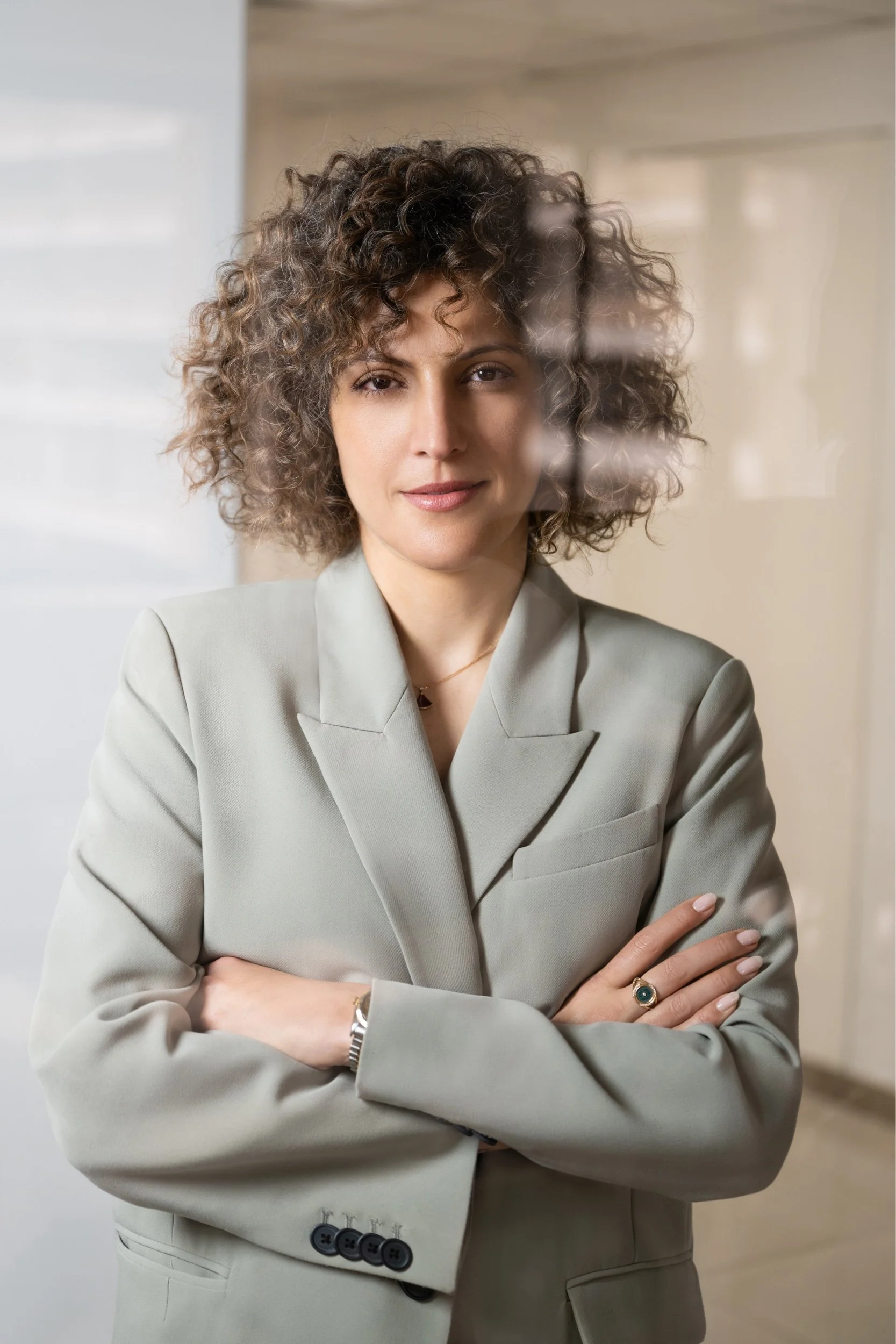 A woman with curly hair wearing a light gray suit, standing with her arms crossed, indoors with a blurred background.