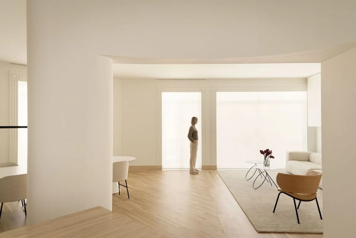 A woman standing in a bright, minimalistic living room with beige furniture, a small glass table with flowers, and large windows with sheer curtains.