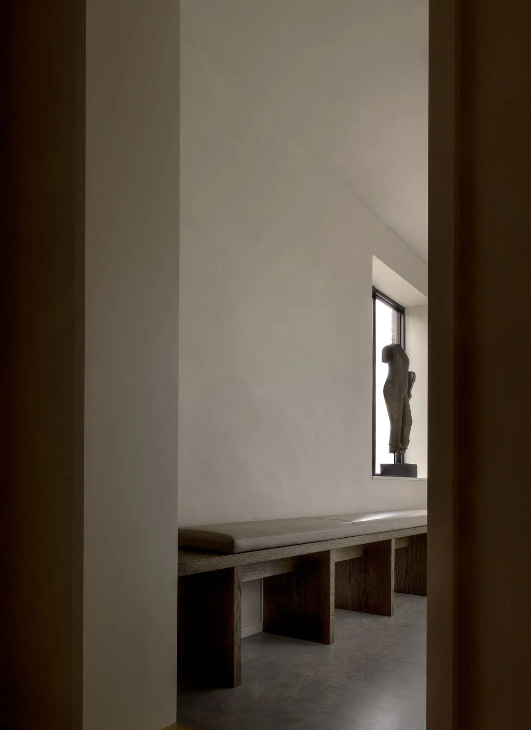 Minimalist interior with a stone sculpture on a window ledge, a bench, and a blank white wall.