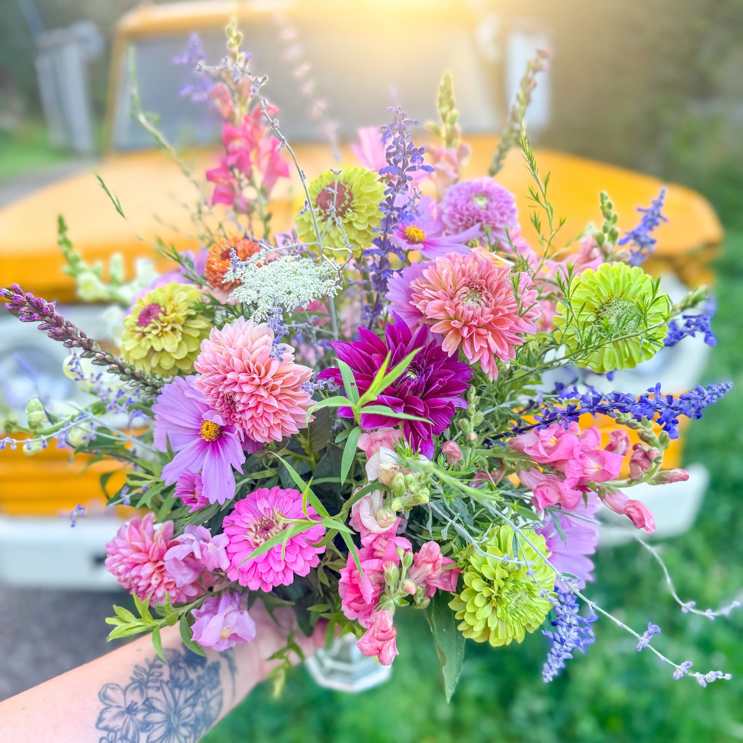 A bouquet of colorful flowers including pink dahlias, purple lavender, yellow-centered green flowers, and white greenish flowers being held by a person with a floral tattoo on their arm, with a blurred green outdoor background and yellow vehicle in the distance.