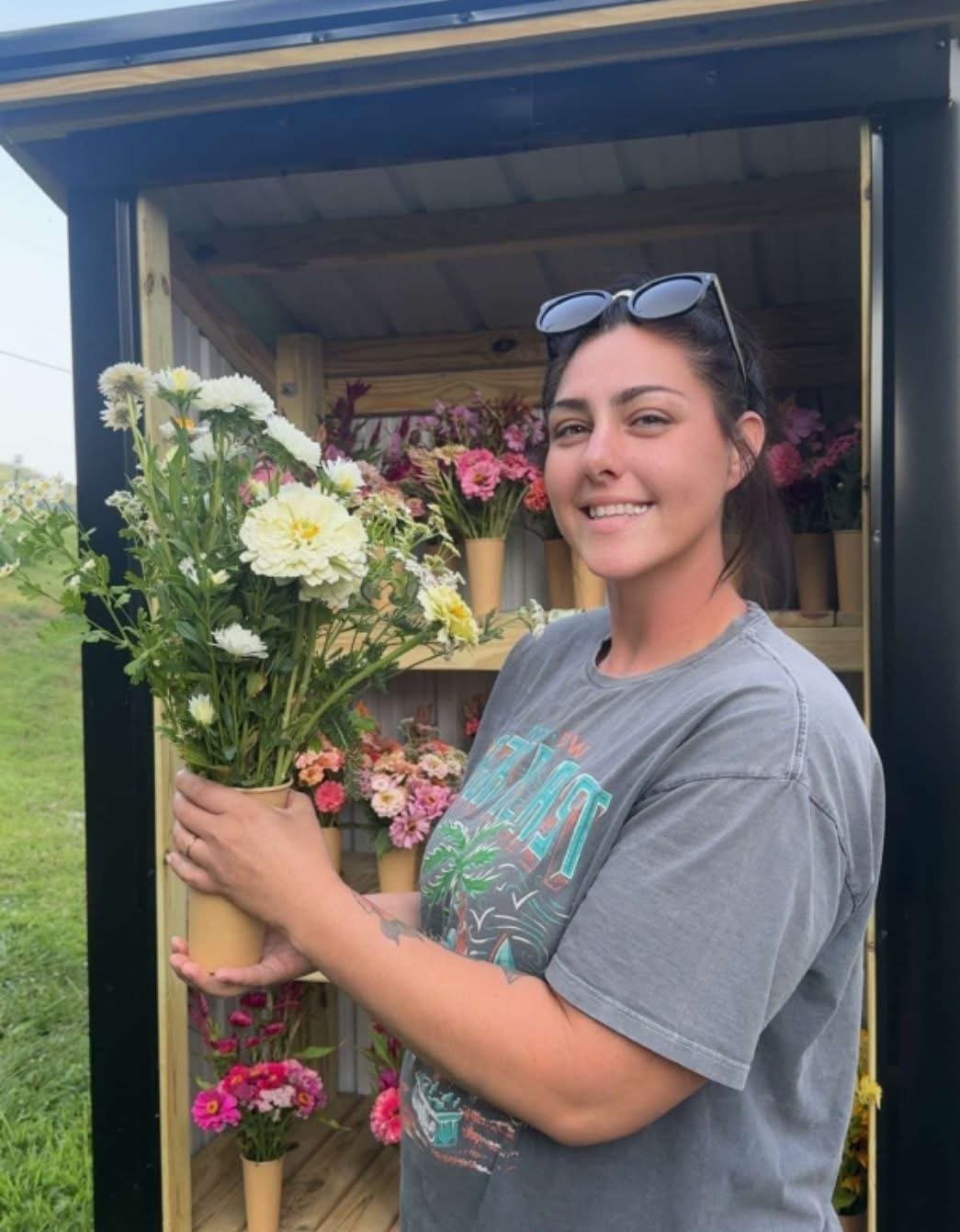 A woman holding a bouquet of white and yellow flowers outdoors, standing in front of a flower stand with various colorful flowers in vases.