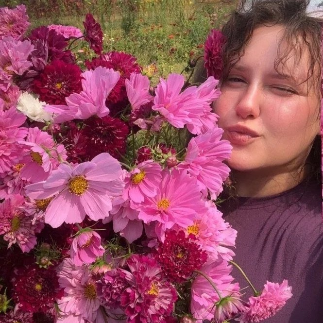 A woman with curly brown hair is taking a selfie beside a large bouquet of pink and red flowers, outdoors in a garden.