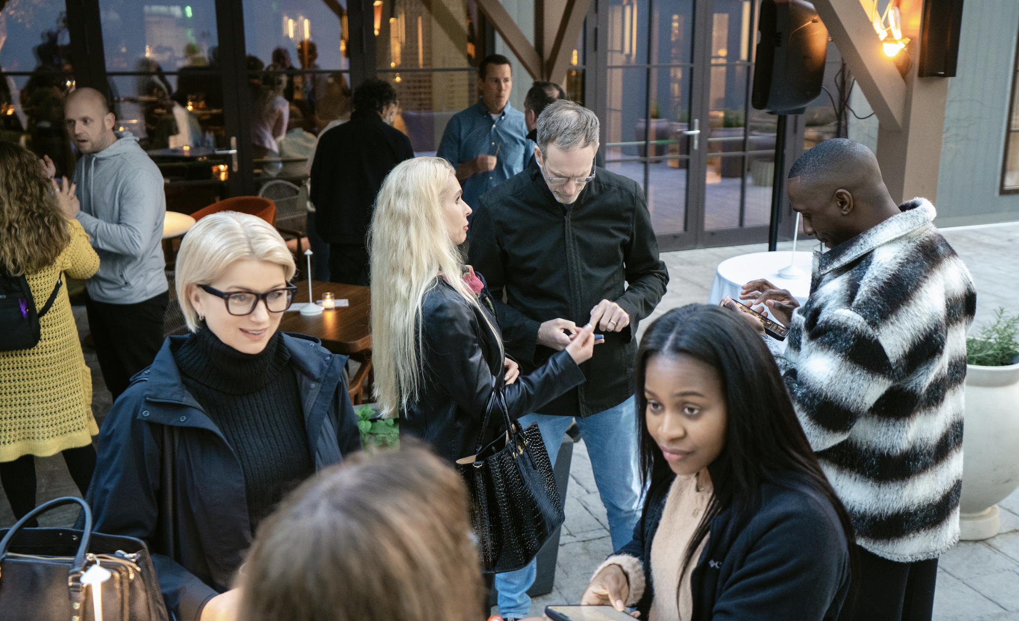 A group of people socializing at an indoor gathering with some sitting and some standing, near large glass doors that lead outside to a patio with more people.