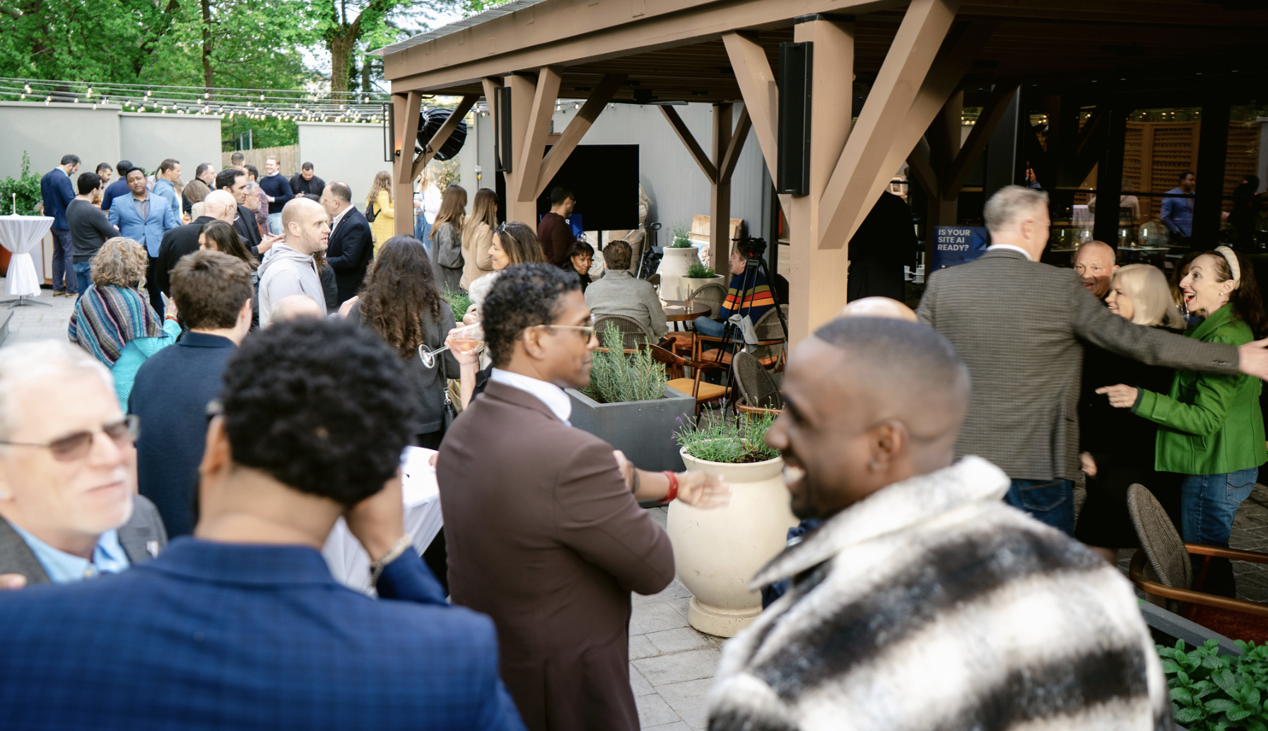 People gathered at an outdoor event, socializing and mingling under a covered patio area, with some seated and some standing, surrounded by trees and outdoor string lights.