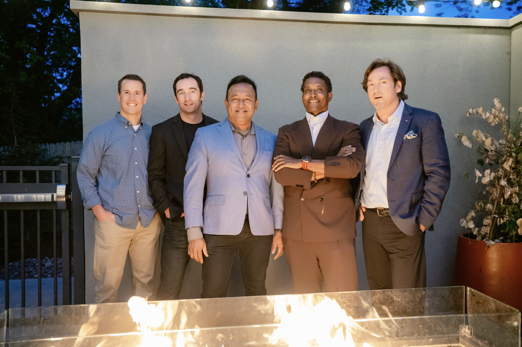 Jay Norris & the Thought Partnr staff dressed in business casual and formal attire standing outdoors at dusk, with a fire pit in the foreground and string lights overhead.