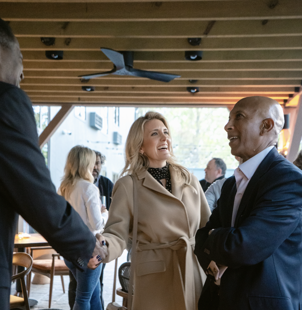 A woman in a beige trench coat shaking hands with a man in a dark suit at a social gathering.
