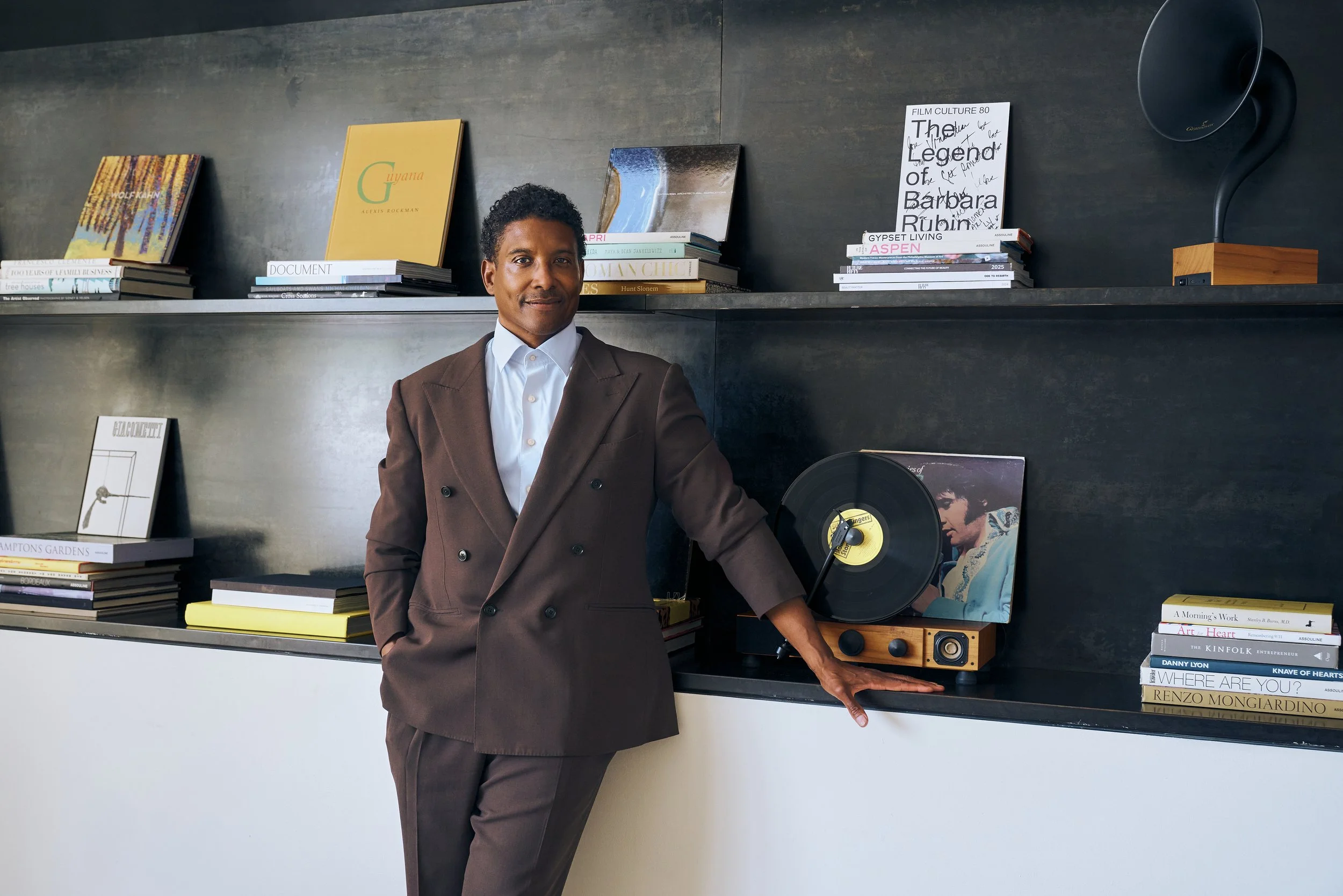 A man in a brown suit and white shirt standing in front of a dark bookshelf displaying books and vinyl records, with his right hand resting on a vintage record player.