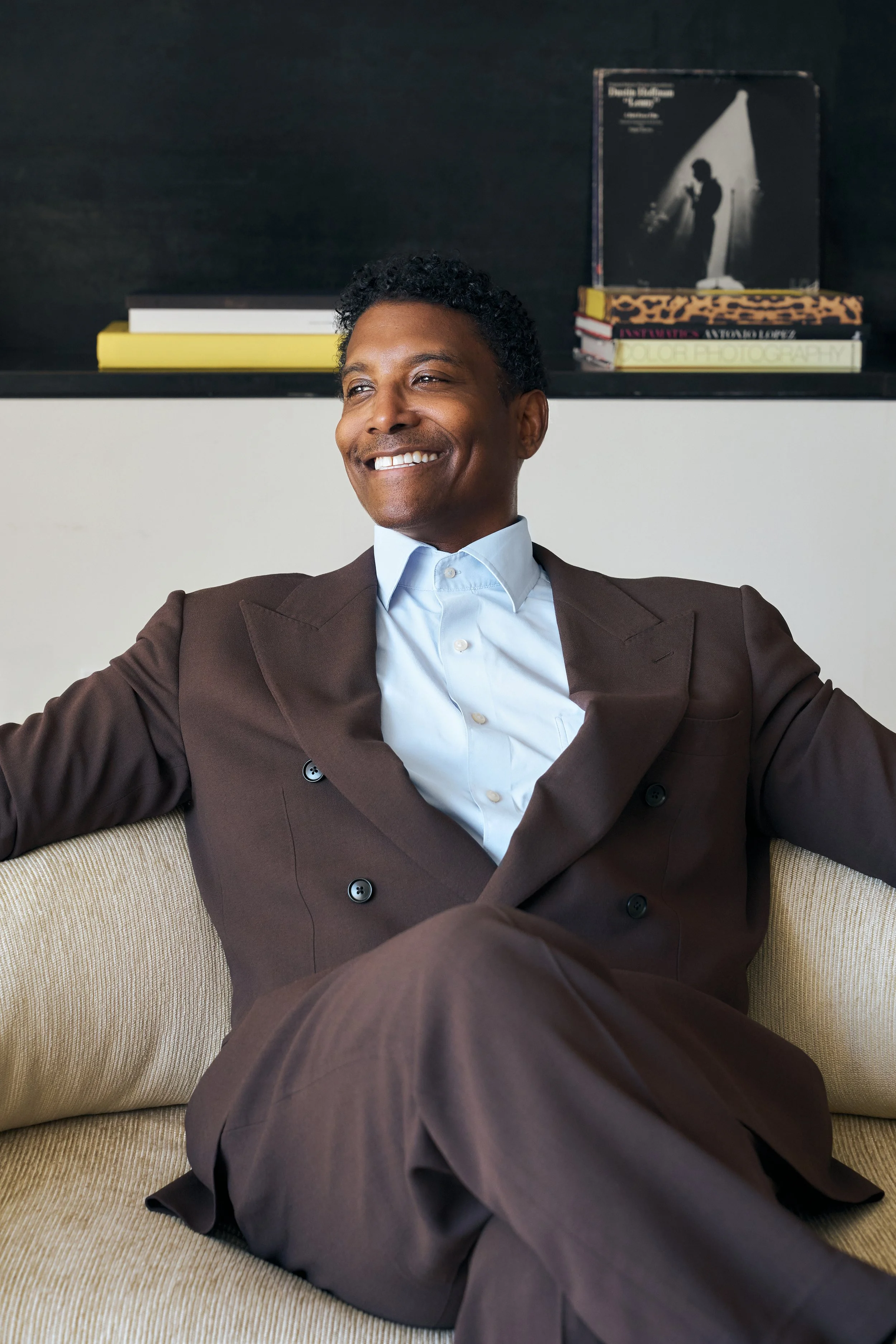 Jay Norris in a brown suit and light blue shirt sitting on a beige couch, with a bookshelf behind him that has several books and a black-and-white photograph of a musician with a guitar in a spotlight.