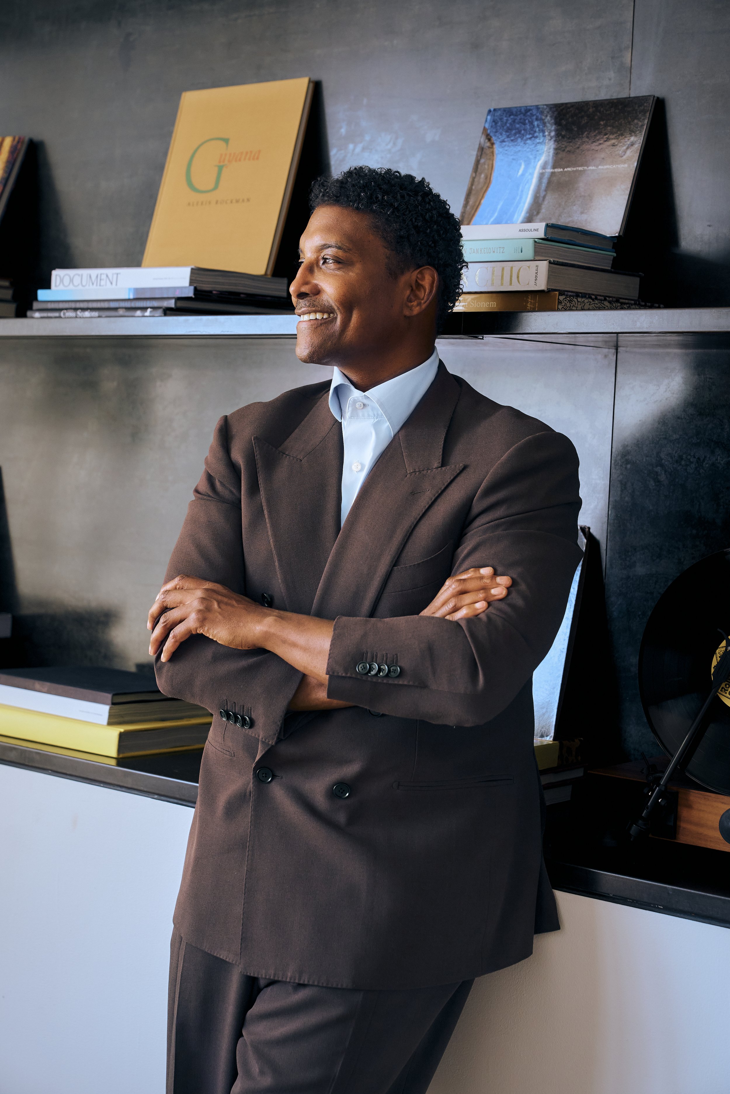 Jay Norris in a suit with arms crossed, smiling, standing in front of a shelf with books and artwork in a modern office setting.
