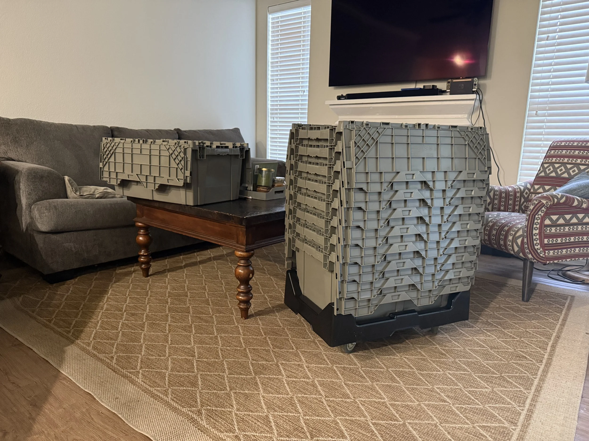 A living room with a gray sofa, wooden coffee table, patterned armchair, and a large plastic crate on wheels filled with smaller crates, all on a beige rug. There are windows with blinds and a wall-mounted TV.
