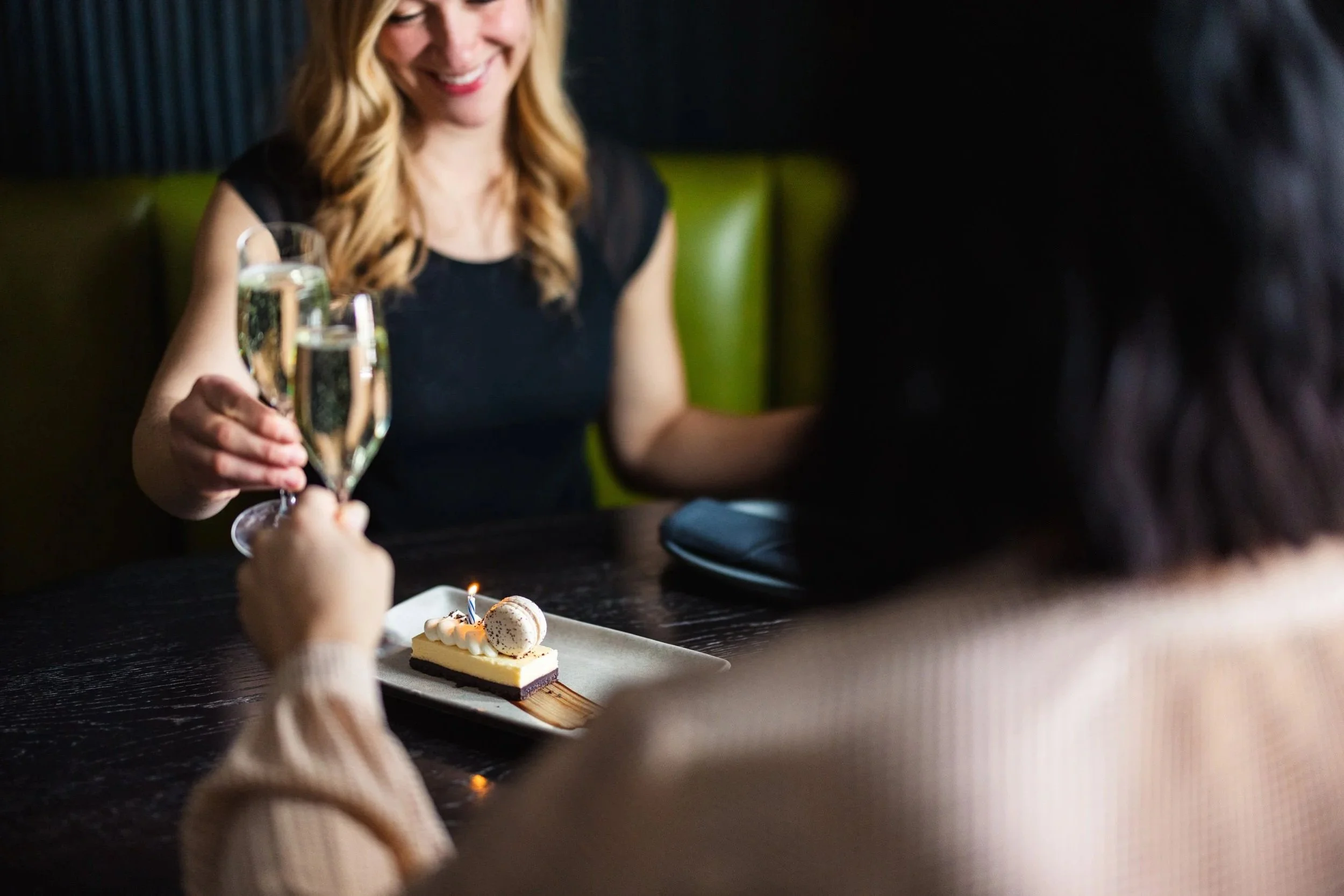 A woman in a black dress celebrating a birthday, holding a glass of champagne, with a small cake with lit candles on a plate in front of her at a restaurant.
