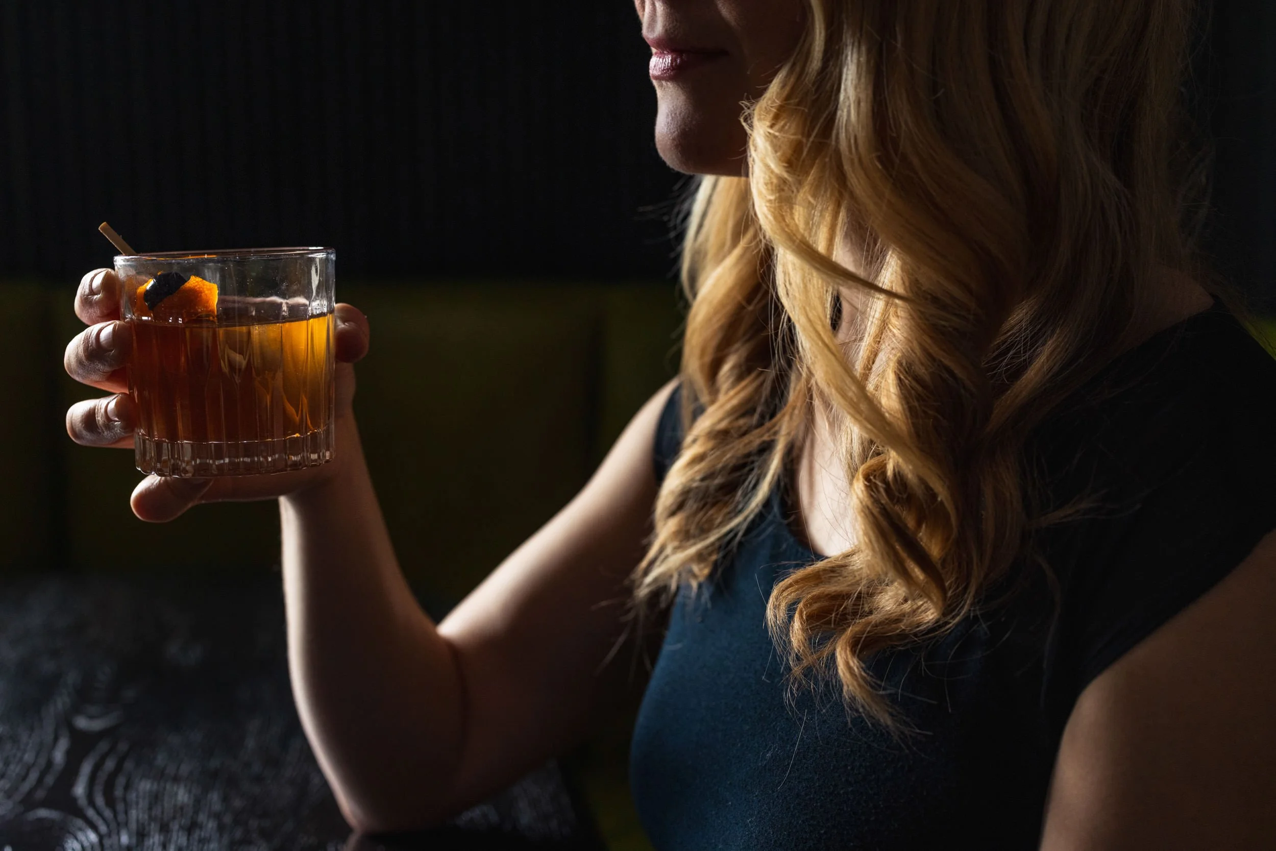 A woman with long, curly blonde hair is sitting in a dimly lit space, holding a glass of amber-colored beverage with a large ice cube and an orange peel garnish. She is wearing a dark-colored top and appears to be smiling softly.