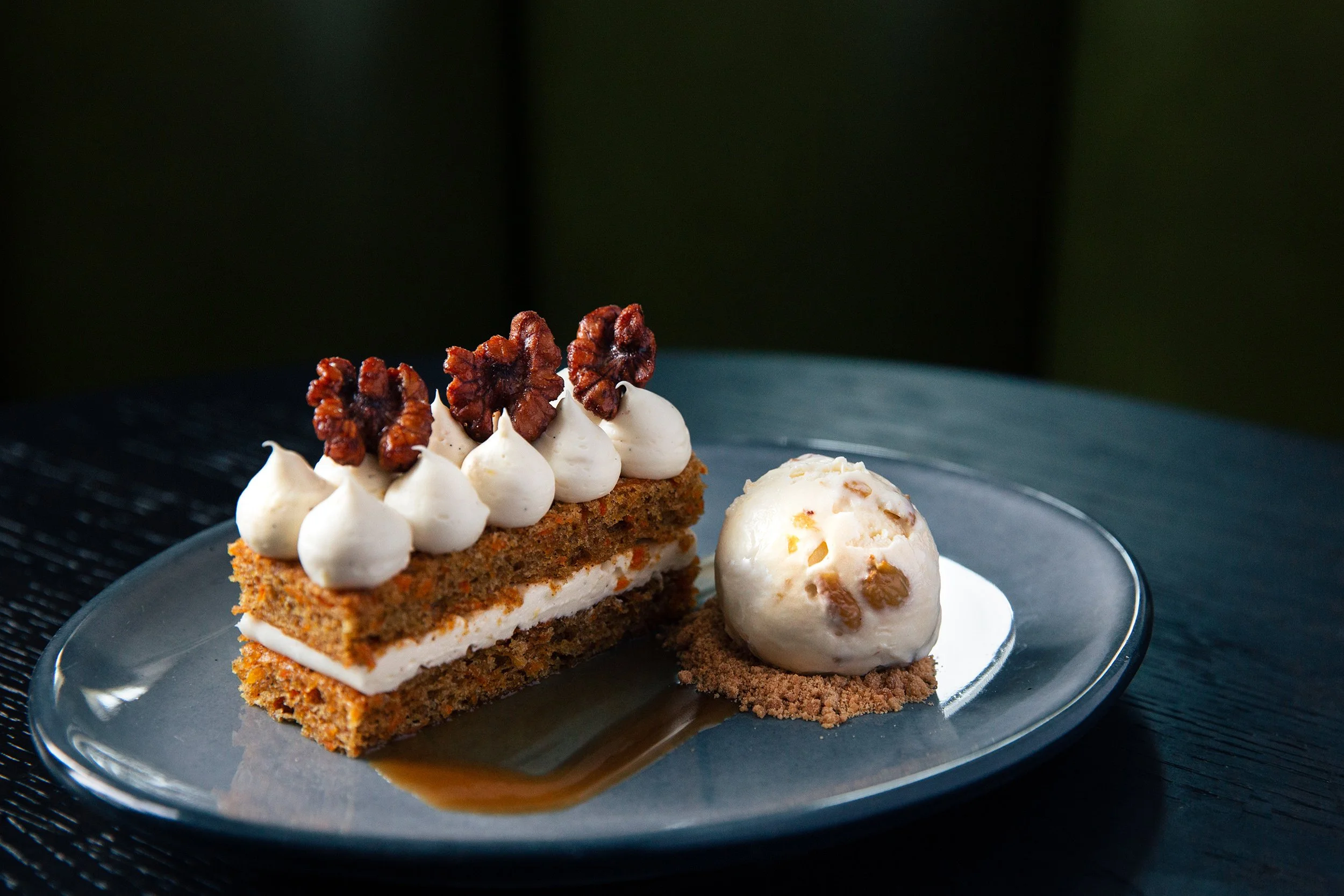 A slice of carrot cake with cream cheese frosting and pecan topping, served with a scoop of vanilla ice cream drizzled with caramel sauce on a black plate.