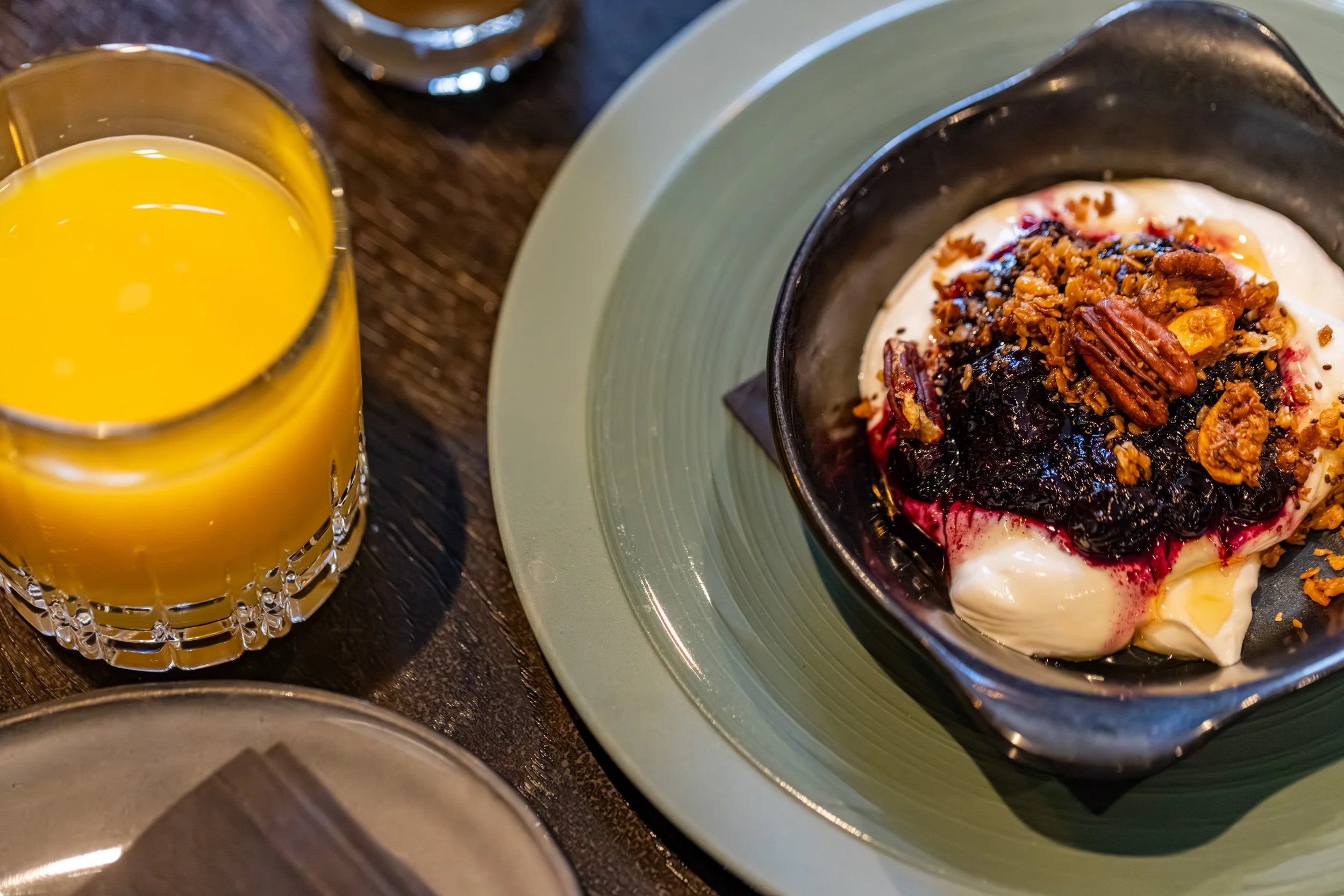 A bowl of yogurt topped with granola, nuts, and berries sits on a green plate. Next to it, a glass of orange juice is partially visible.