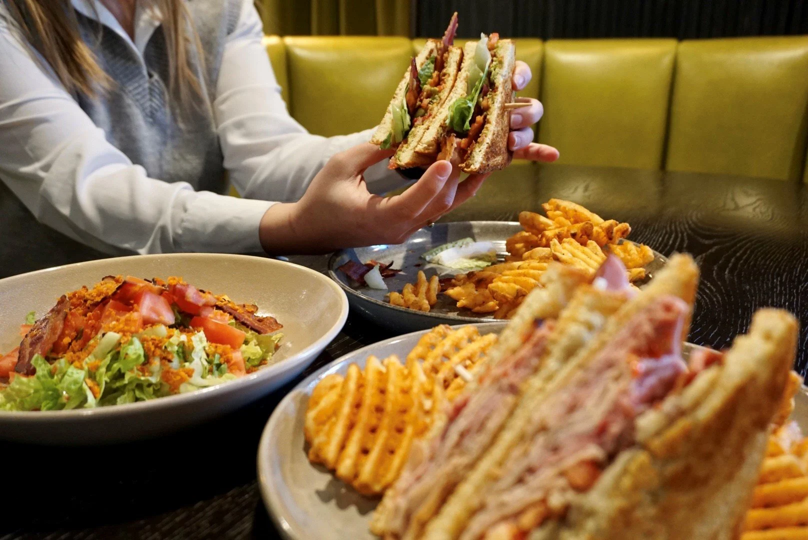 Person holding a triple-decker club sandwich with fries and a salad on a black dining table in a restaurant with green seating in the background.