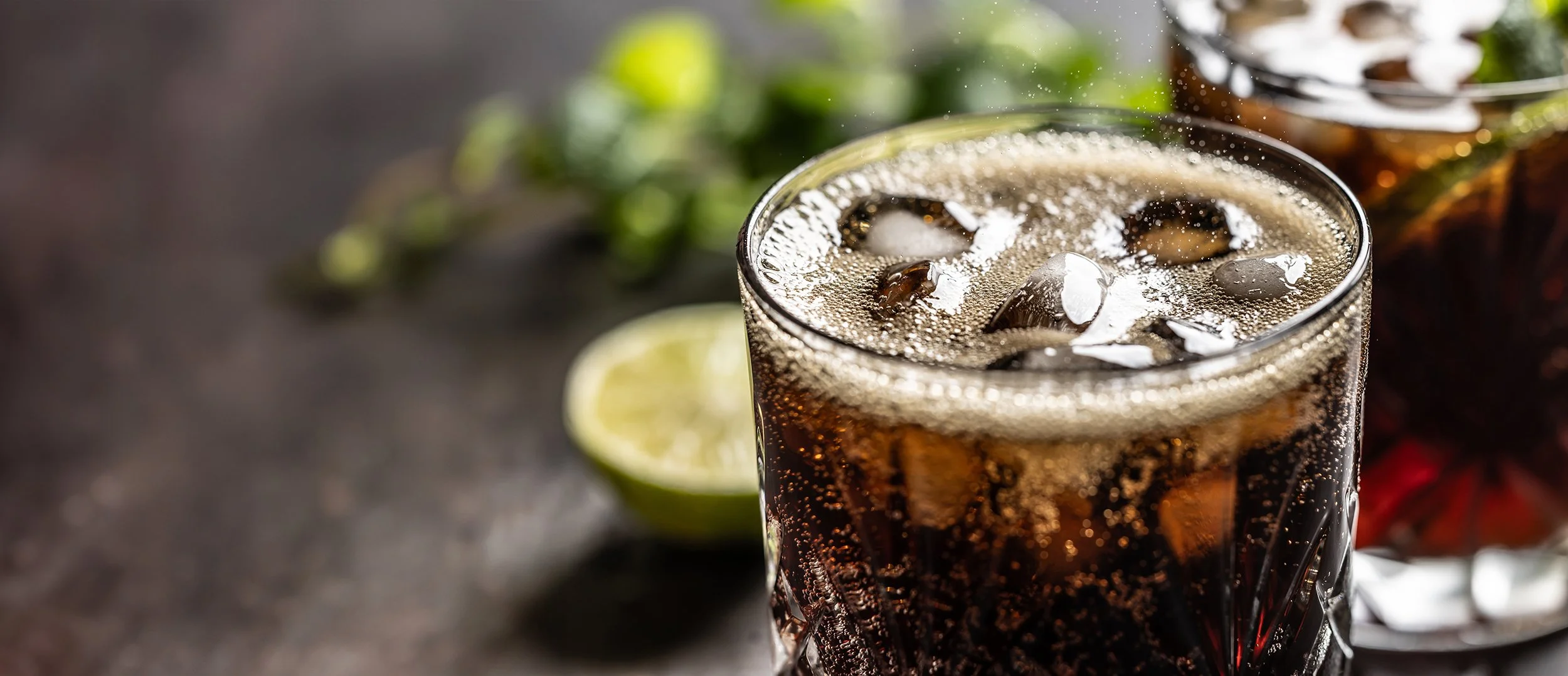 A glass of dark soda with ice cubes, bubbles, and condensation on the outside, placed on a dark surface with lime slices and green herbs in the background.