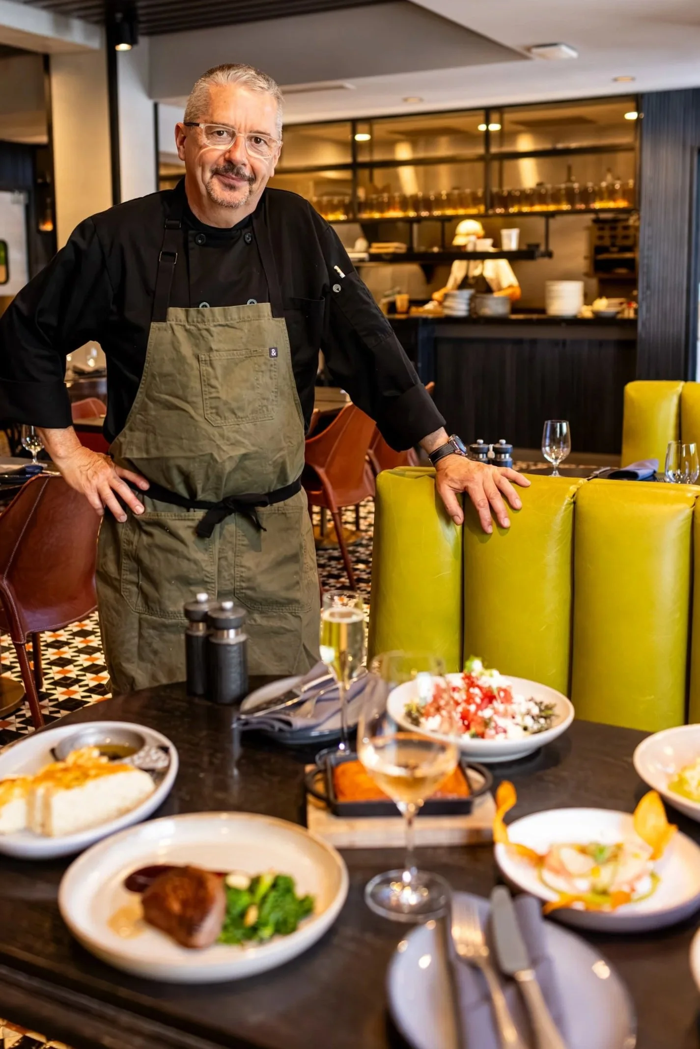 A smiling chef in a black shirt and green apron stands behind a dining table with various dishes. The background shows a modern restaurant kitchen with shelves and staff working.