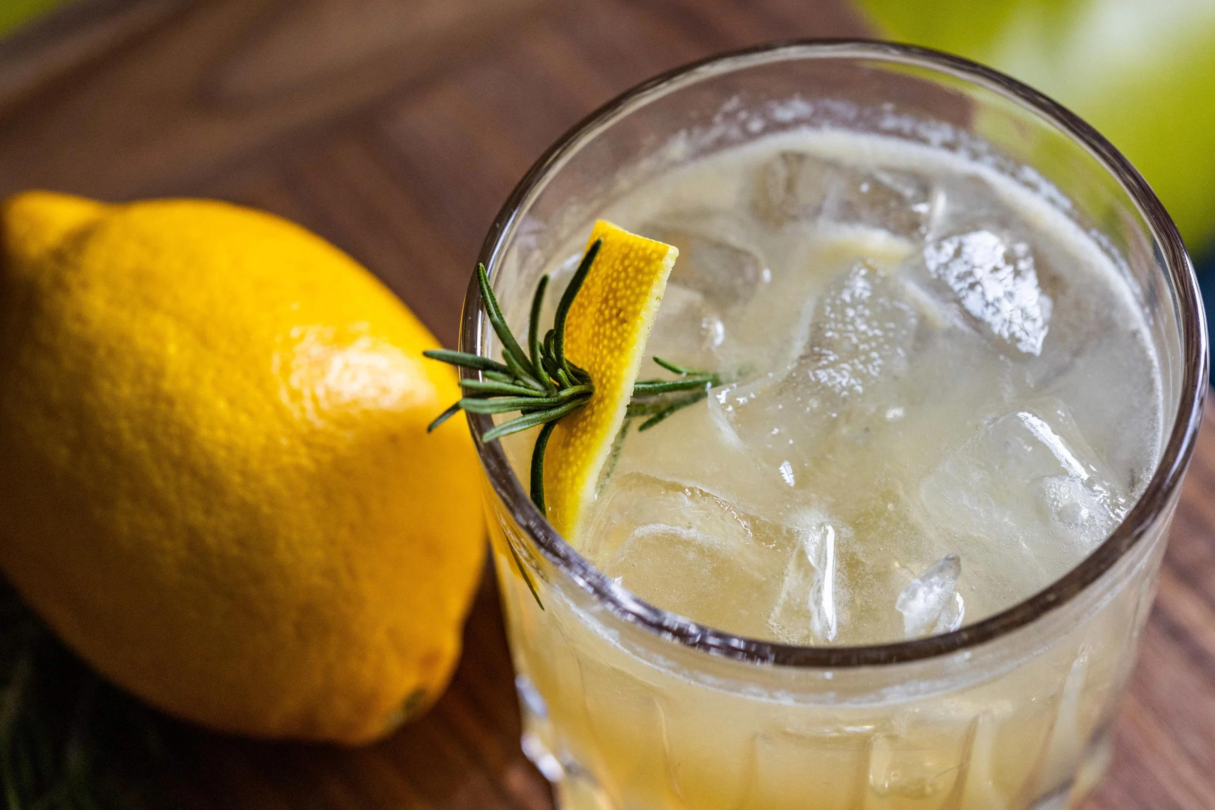 A glass of lemonade garnished with a lemon peel and sprig of rosemary beside an uncut lemon on a wooden surface.