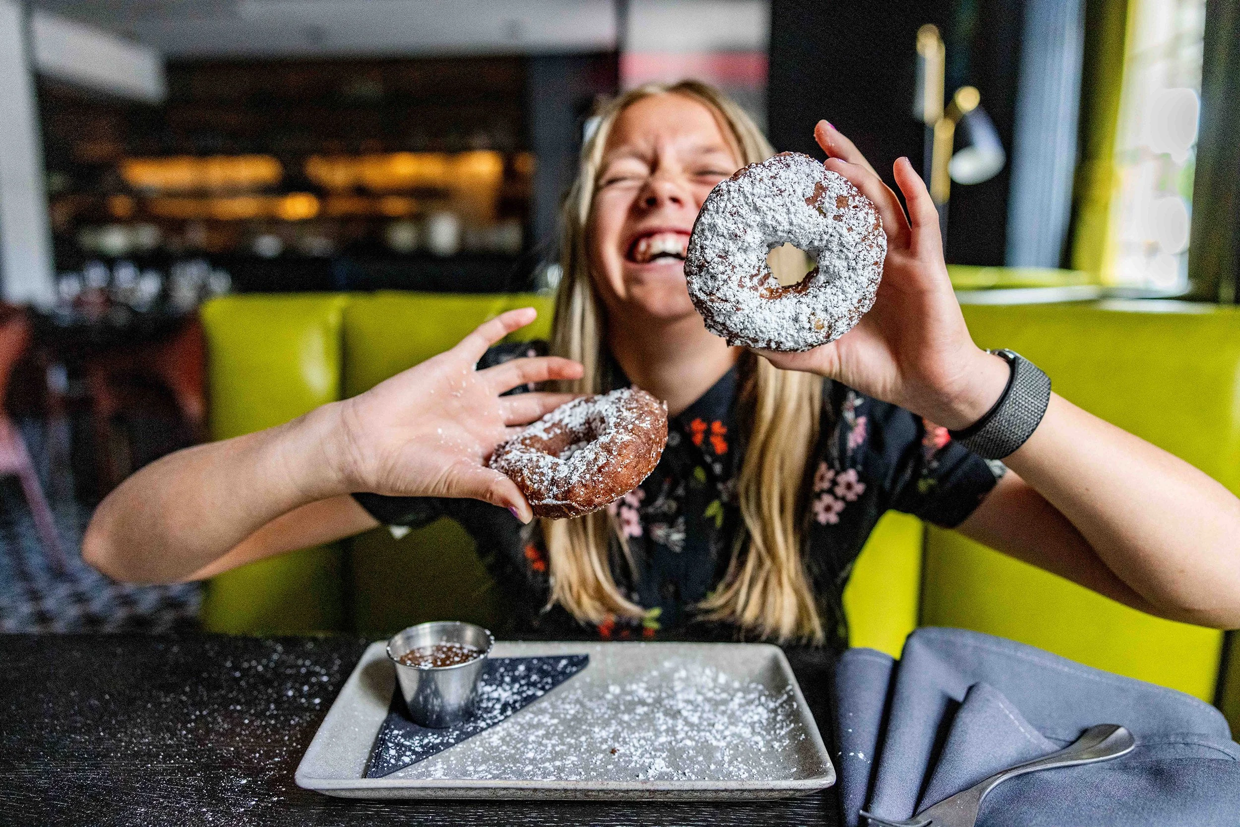 A woman with long blonde hair smiling and holding up a powdered sugar-covered donut. She is sitting at a table with a tray of donuts and a small cup of chocolate sauce, in a brightly lit restaurant.