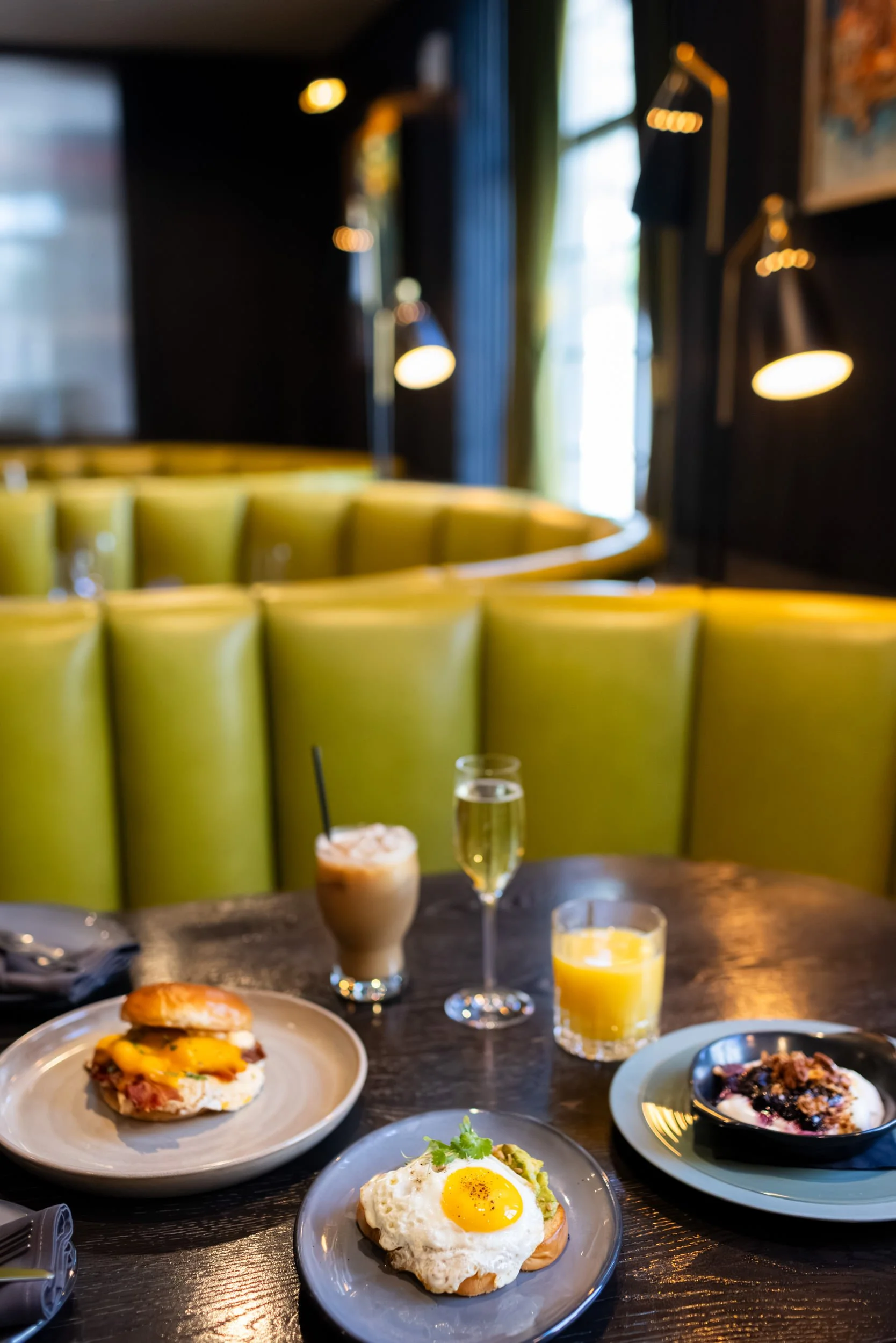 A breakfast table with a plate of eggs and toast, glasses of orange juice and champagne, and a smoothie, in a cozy restaurant with green upholstered booths and warm lighting.
