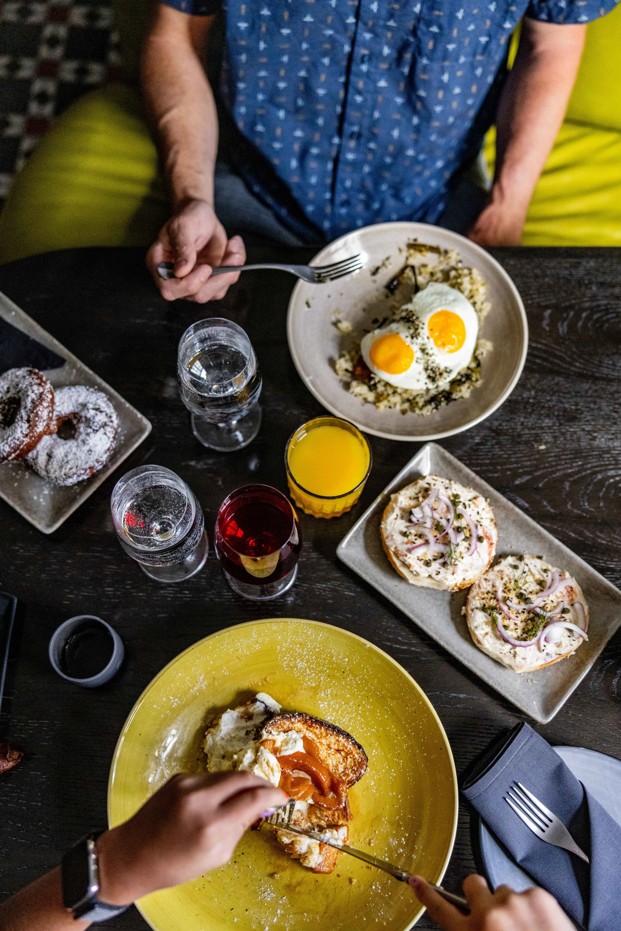 A person is seated at a table with various food dishes. The person is holding a fork and knife, cutting into a dessert on a yellow plate, which appears to be a slice of cake with whipped cream and caramel. On the table are glasses of water and other drinks, a plate with two fried eggs with black seasoning, and a rectangular platter of open-faced sandwiches with various toppings like onions and herbs.