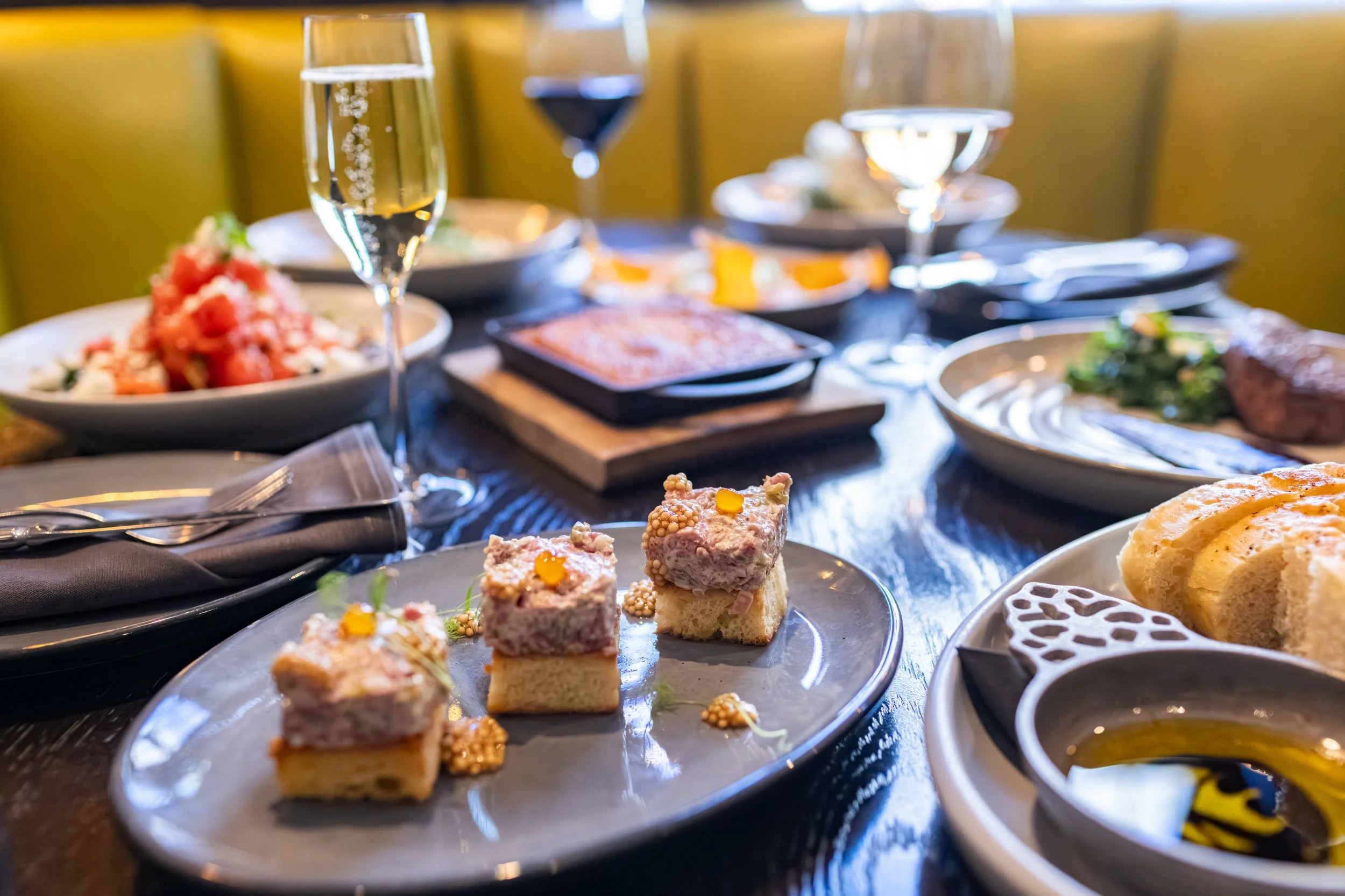 A table with various dishes including small bites, desserts, and glasses of sparkling and red wine. The focus is on small, layered sweet treats with toppings.