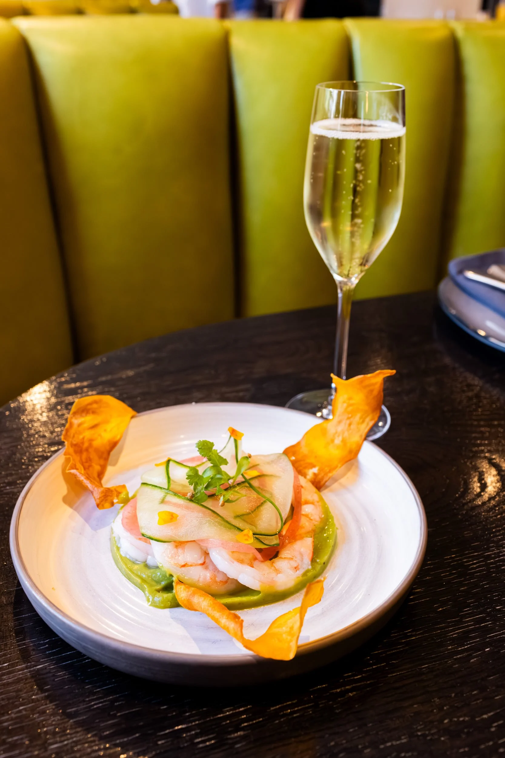 A plate of shrimp ceviche garnished with fresh herbs and served with crispy plantain chips. A glass of sparkling beverage is in the background.