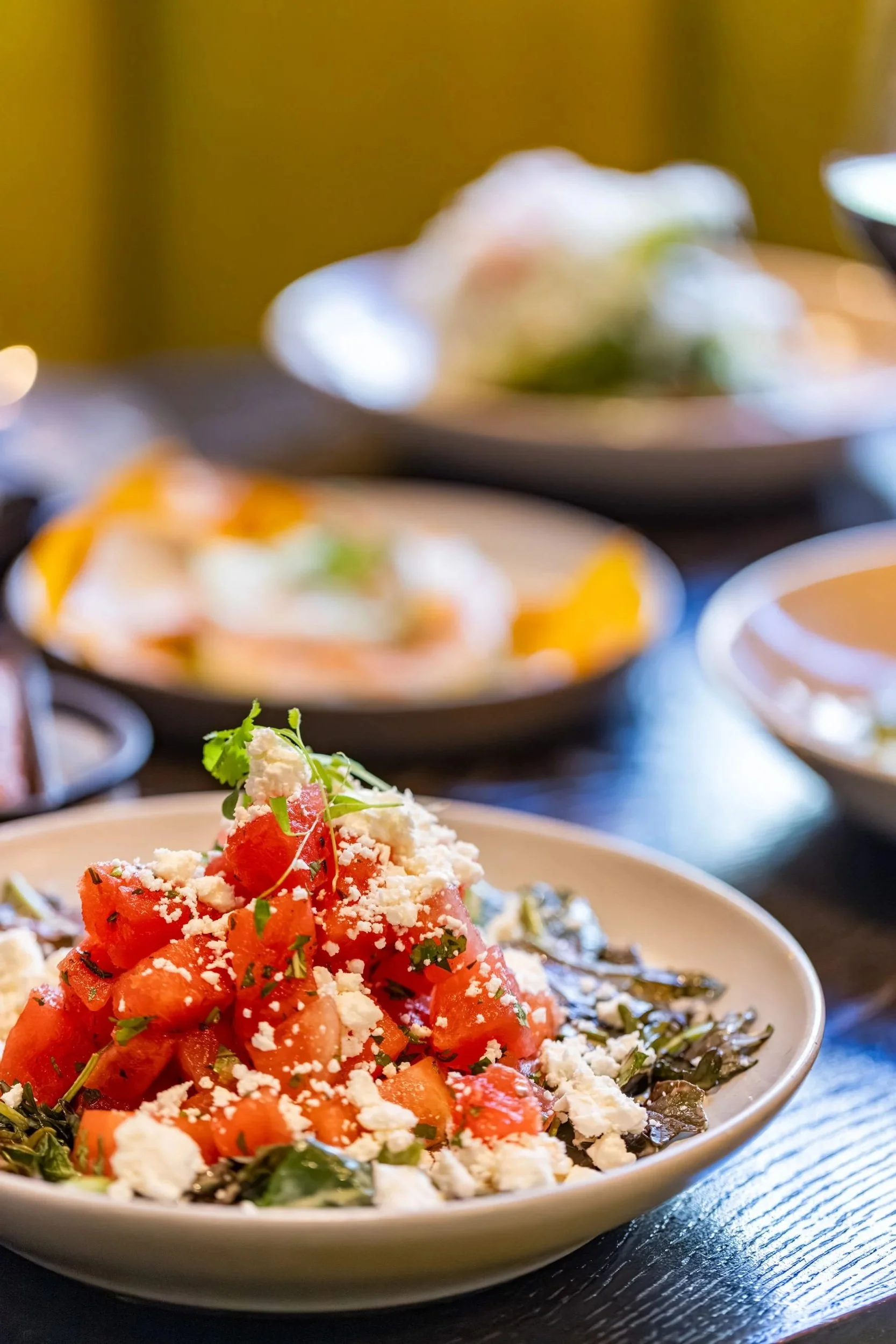 Close-up of a plate of diced tomatoes, topped with crumbled cheese and chopped herbs, with a variety of other dishes blurred in the background.