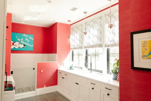 Bright kitchen with red painted walls, white cabinets, large windows with patterned blinds, hanging pendant lights, and framed artwork on the walls.