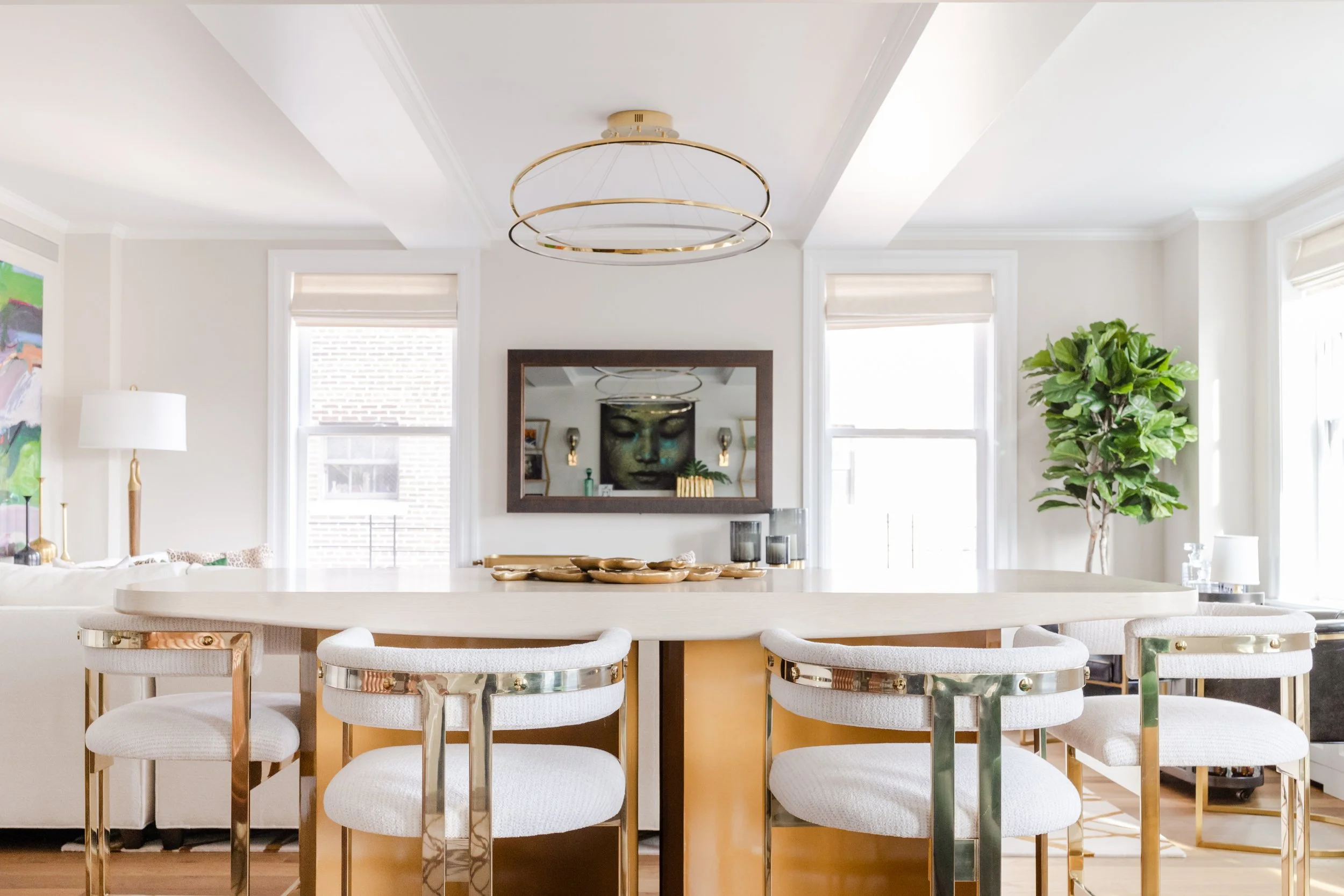 A dining room with a white table, six white chairs with gold accents, a large mirror on the wall, decorative items on the table, a modern chandelier, and a large green leafy plant near the window.