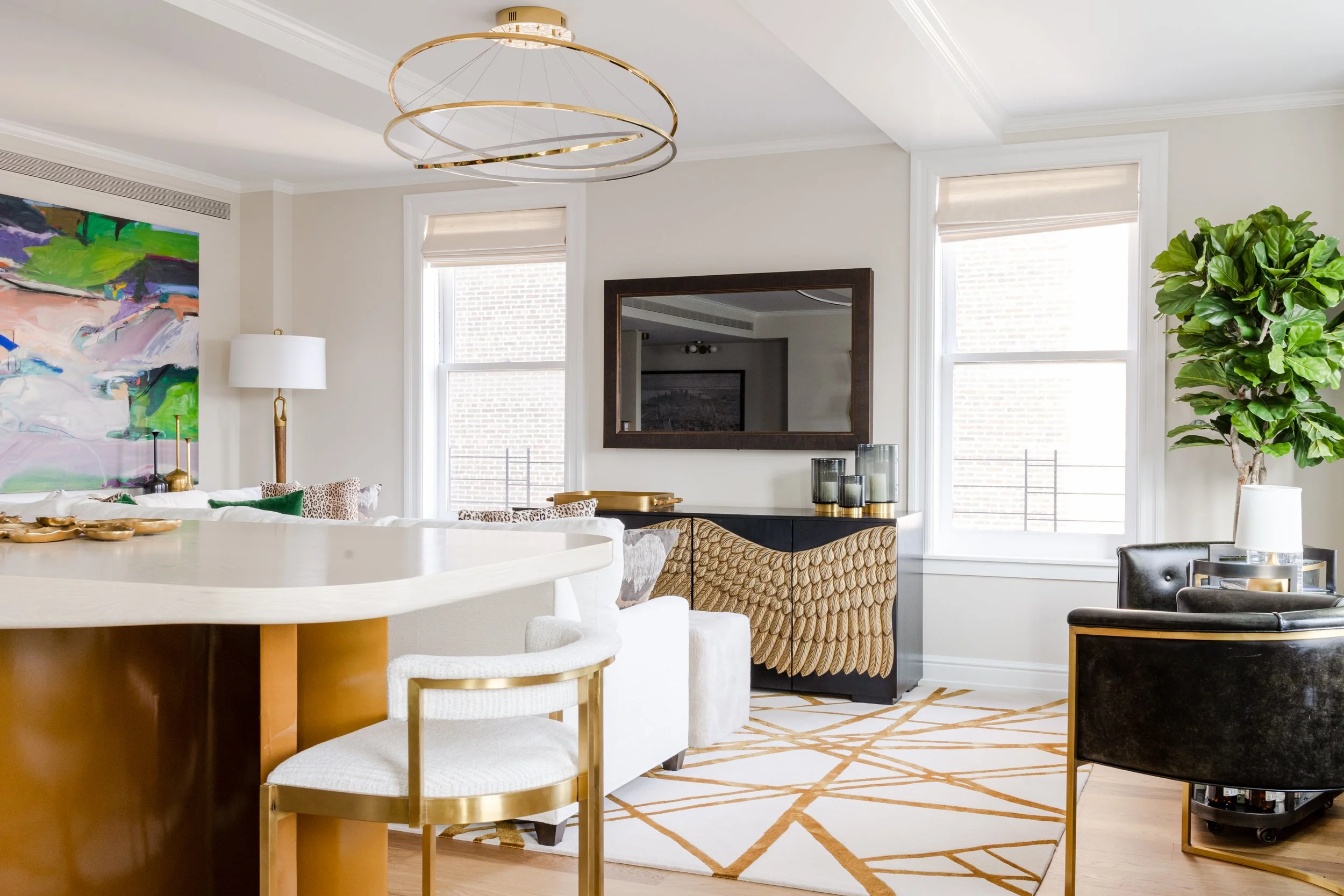 A modern living room with white walls, large windows, and contemporary furniture. There is a colorful abstract painting on the wall, a tall floor lamp, a black sideboard with decorative candles, and a large leafy plant. The room features a gold and w