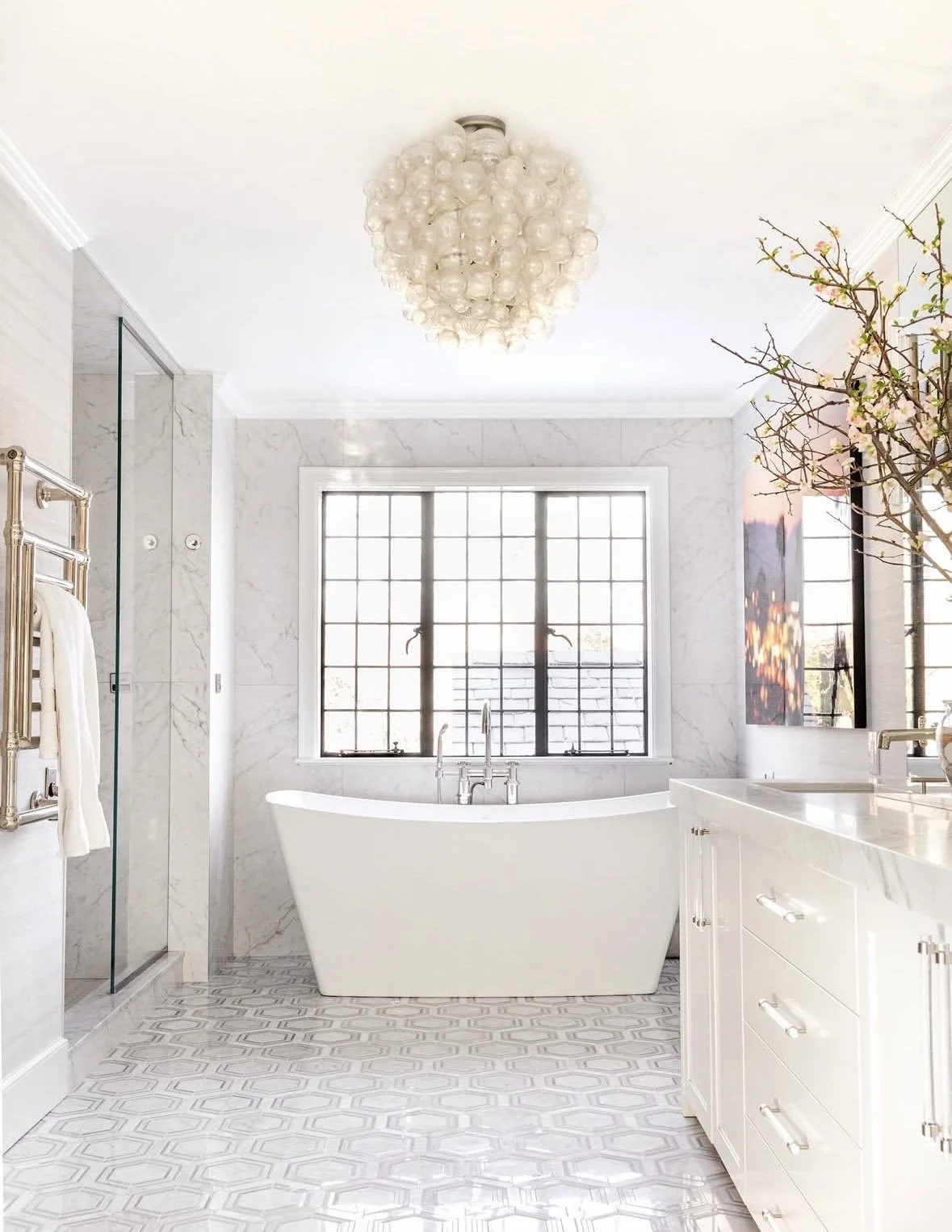 Bright, modern bathroom with a freestanding bathtub, large window, white marble walls, patterned tile floor, chandeliers, and decorative elements.