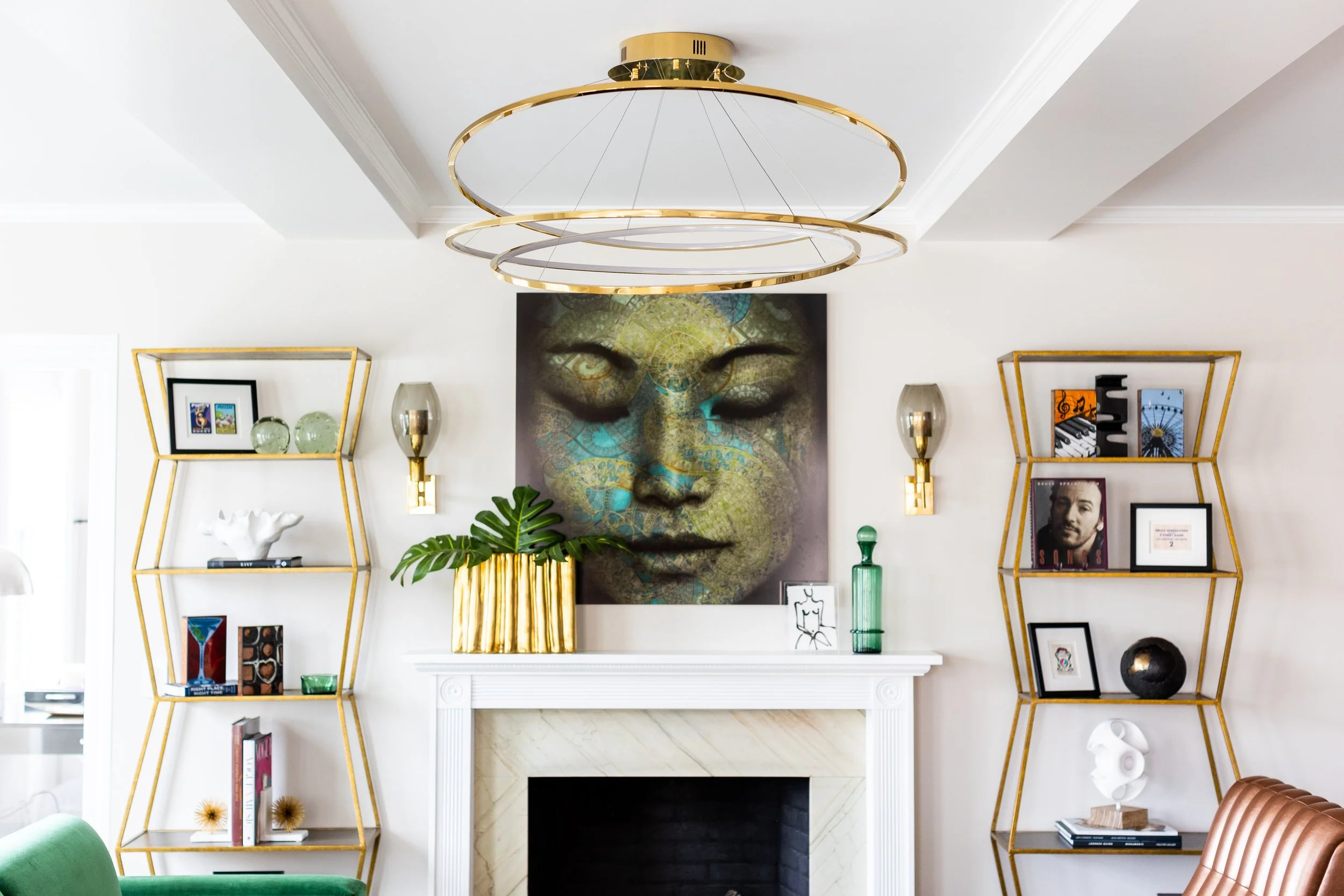 Living room with white fireplace, gold geometric shelves on either side, contemporary artwork of a face above the fireplace, and various decorative objects including vases, books, and framed pictures.