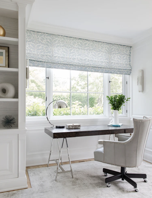 A home office with a large window, a wooden desk, a white office chair, a lamp, a potted plant, and decorative items on the shelves.