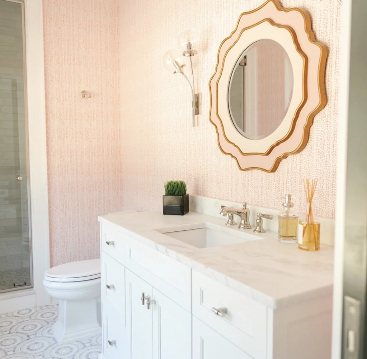 A bathroom with a white vanity, marble countertop, rectangular undermount sink, silver fixtures, a decorative mirror, a small potted plant, reed diffuser, and a wall-mounted light fixture, with a pink textured wall and a shower door visible on the le
