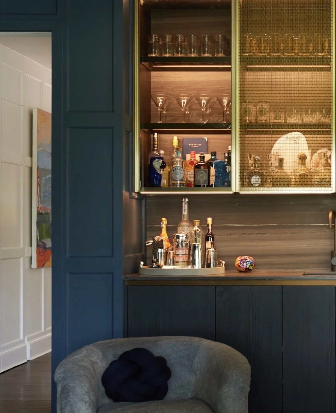 A home bar with a blue wooden cabinet, shelves with various liquor bottles, glasses, and bar tools. A colorful decorative item is on the counter, with a dark gray chair in the foreground.