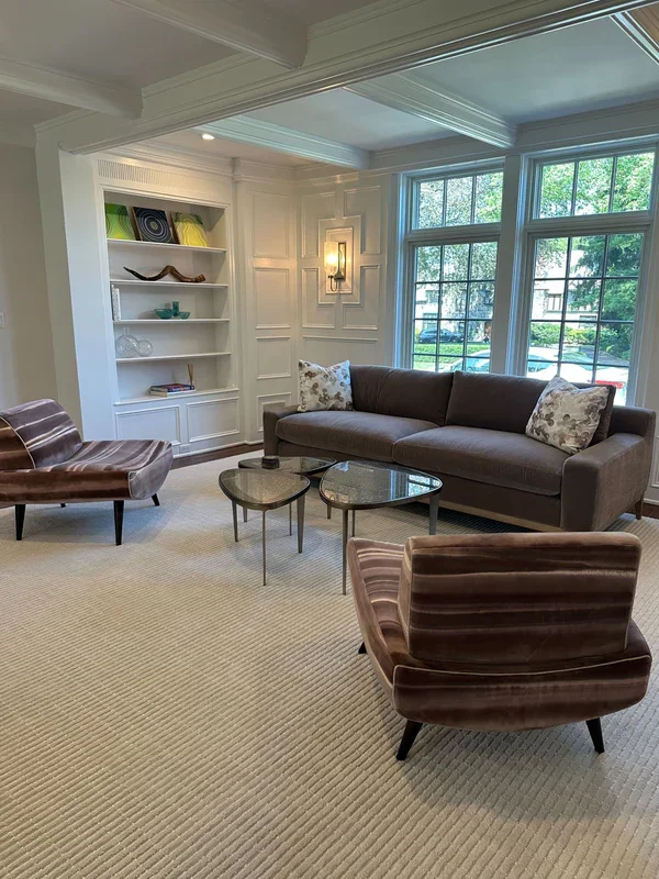 Living room with a gray sofa, two striped armchairs, a built-in bookshelf, large windows, and a beige carpet.