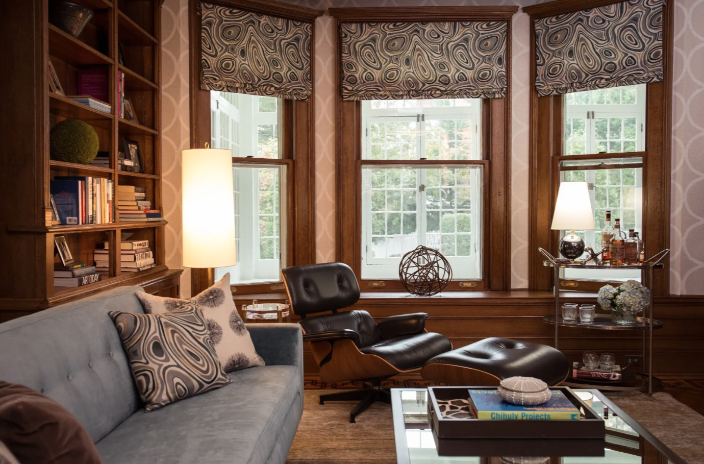 A cozy living room with large windows, a gray sofa with patterned cushions, a black leather lounge chair, wooden shelving filled with books, and a side cart with lamps, bottles, glasses, and flowers.