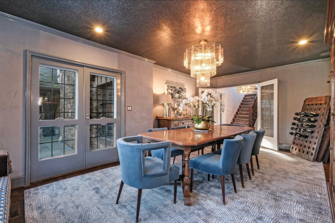 A dining room with a large wooden table, eight blue upholstered chairs, a chandelier, a sideboard with flowers, and glass double doors leading outside.