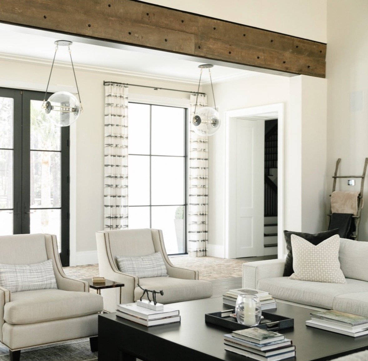 Living room with white sofas, a black coffee table with books, decorative items, large windows with patterned curtains, and a wooden beam on the ceiling.
