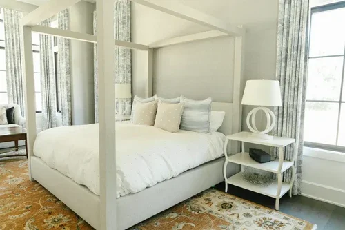 A cozy bedroom with a white canopy bed, decorative pillows, a white side table with a lamp, and a large window with curtains.