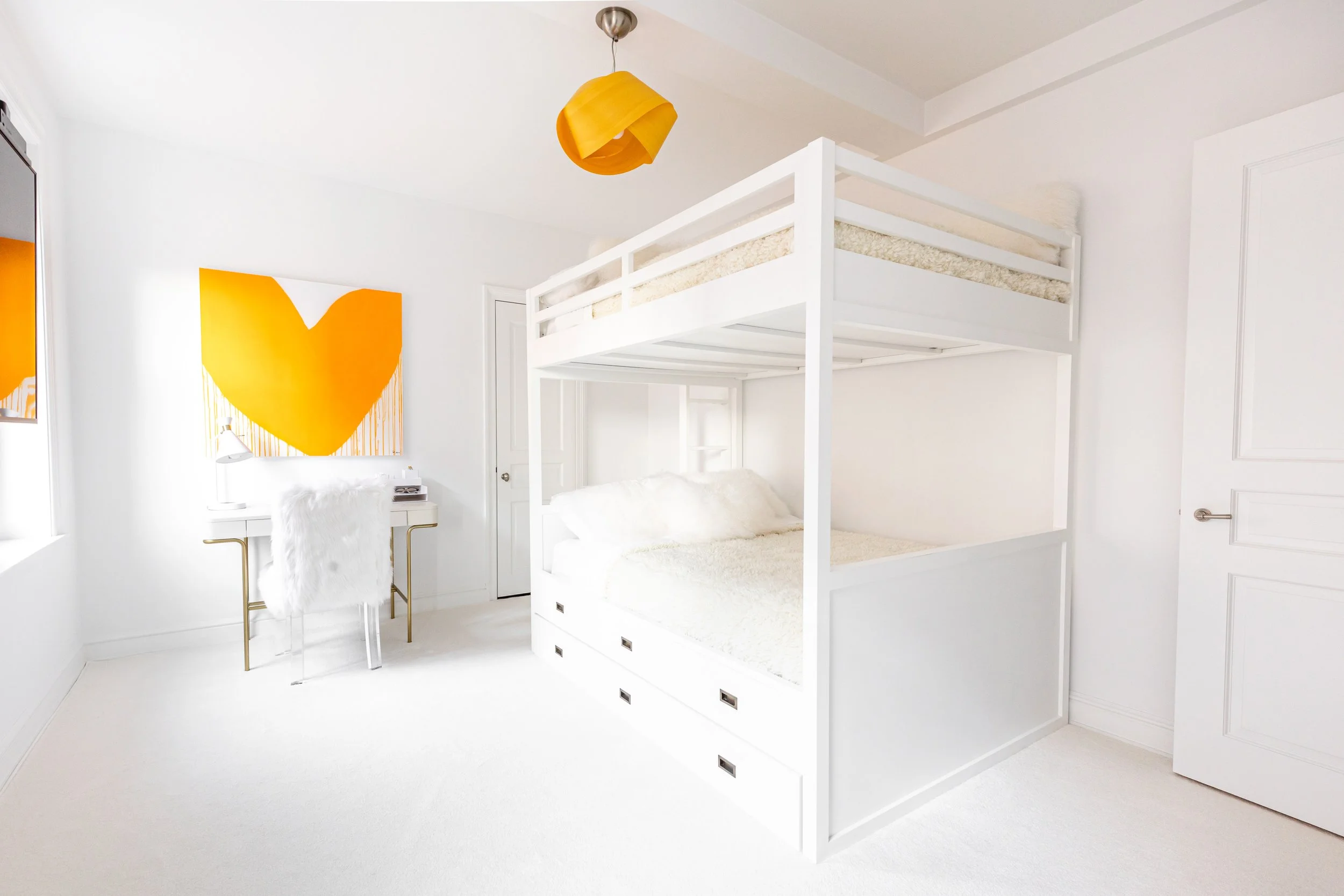 A white bedroom with a white bunk bed, a white fluffy chair, a small white desk with a lamp, a large yellow heart painting, and a yellow lampshade hanging from the ceiling.