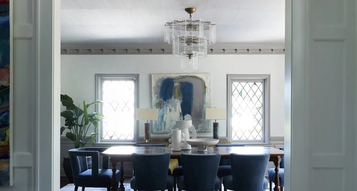 Dining room with a large wooden table, blue chairs, two windows with diamond-patterned panes, a large abstract painting, and a chandelier hanging from the ceiling.