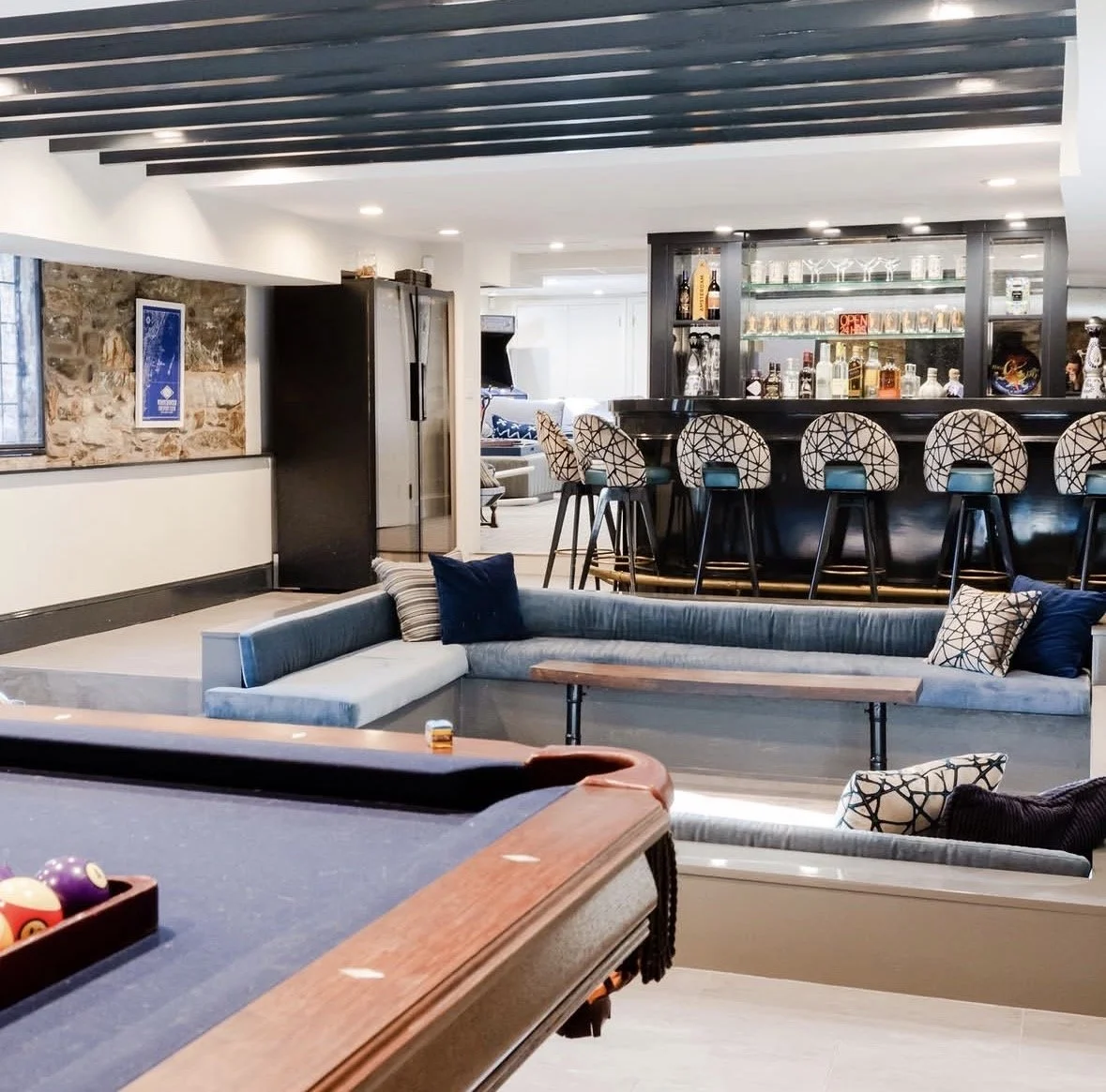A cozy lounge with a bar area featuring high chairs, a pool table, a sectional sofa, and cushions, with a stone wall and modern decor.