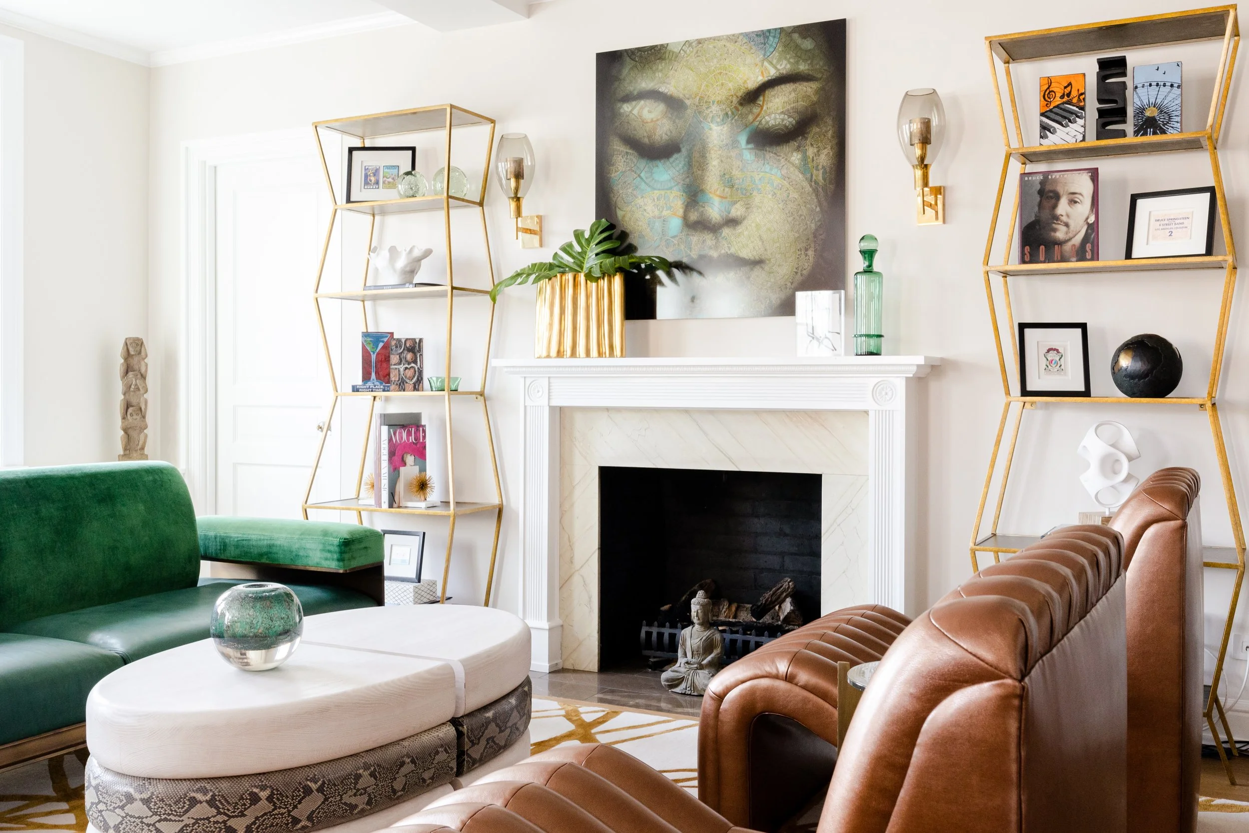 Living room with a white fireplace, green velvet sofa, tan leather sofa, modern decor, abstract face painting on the wall, and gold-framed shelves with decorative items and framed pictures.