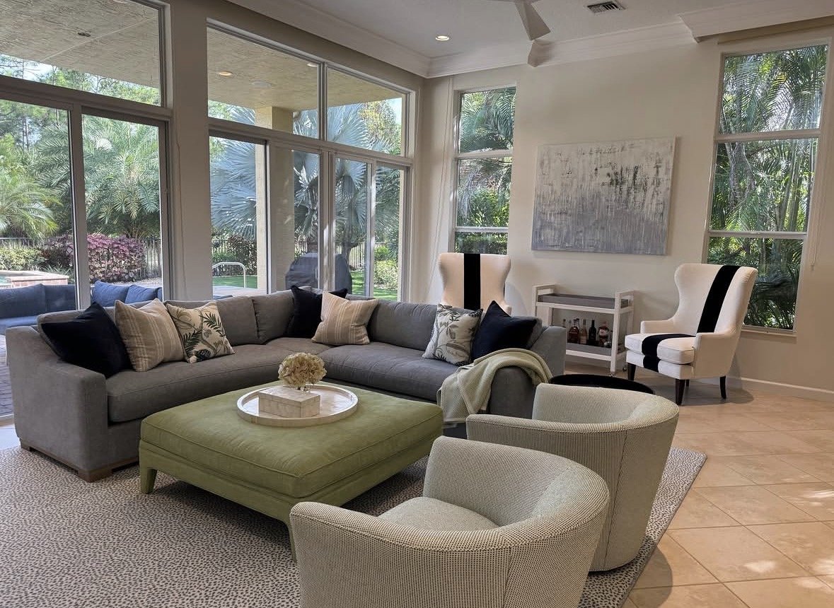 Bright living room with large windows showing outdoor greenery, beige sofa with decorative pillows, two white armchairs with black accents, a green ottoman, and a coffee table with flowers, a textured rug, and artwork on the wall.