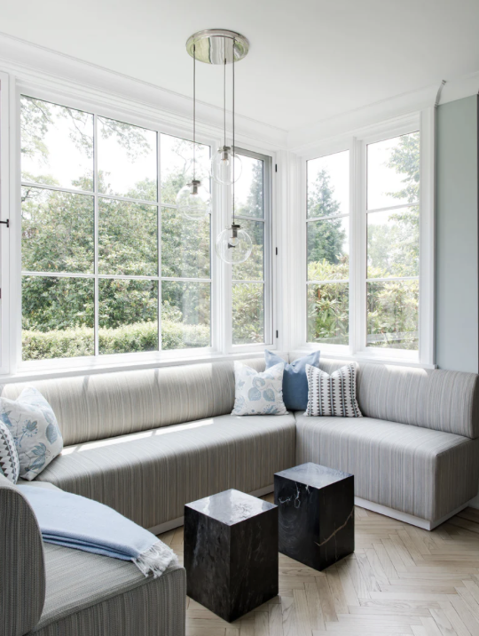 Bright sunlit living room corner with large windows, a beige striped sectional sofa with decorative pillows, two black marble side tables, and a modern hanging light fixture with glass globes.